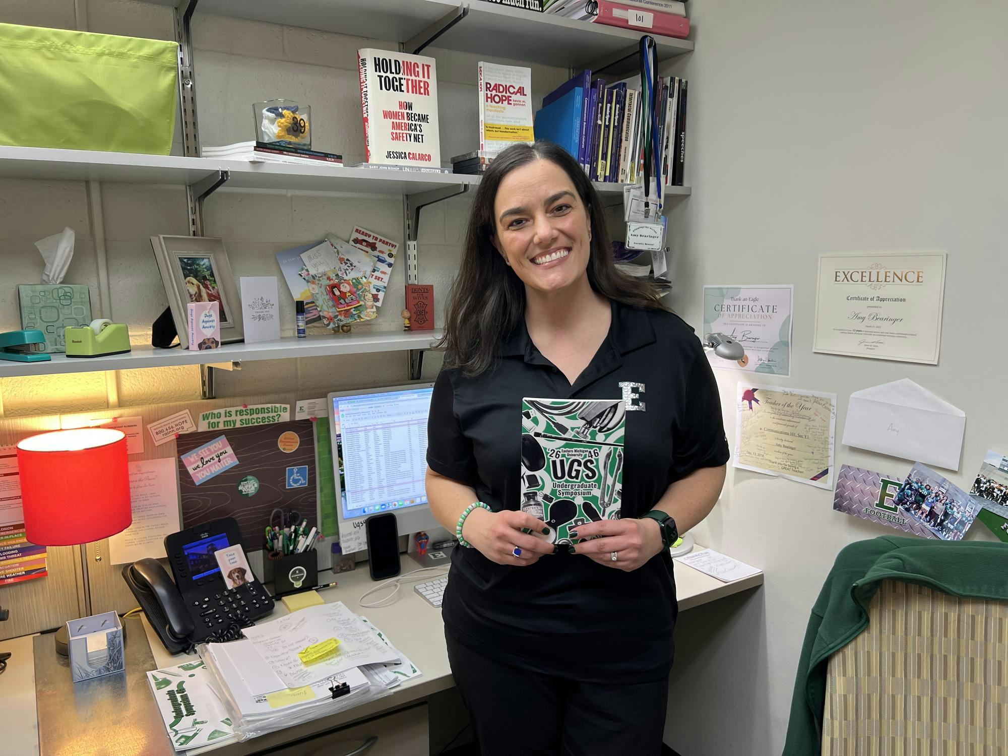 A woman wearing a block E shirt stands in front of a desk area with multiple books, certificates and stickers. She is holding a green, black and white book, which has a collage-style cover.