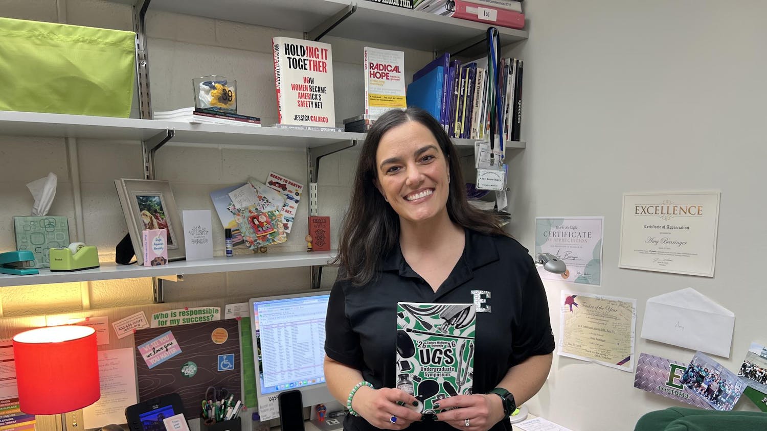 A woman wearing a block E shirt stands in front of a desk area with multiple books, certificates and stickers. She is holding a green, black and white book, which has a collage-style cover.