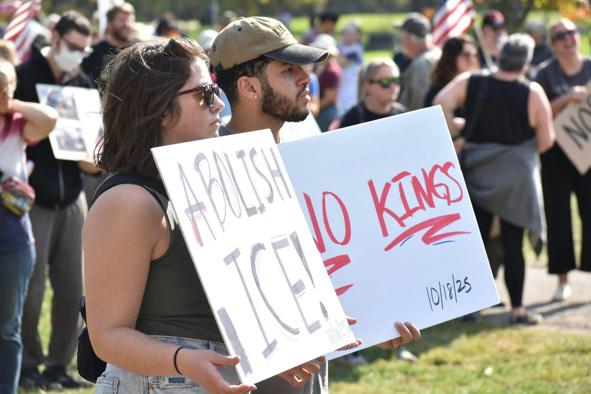 Two people hold white signs that read "ABOLISH ICE," and "NO KINGS," outdoors, surrounded by other people holding signs and waving American flags.