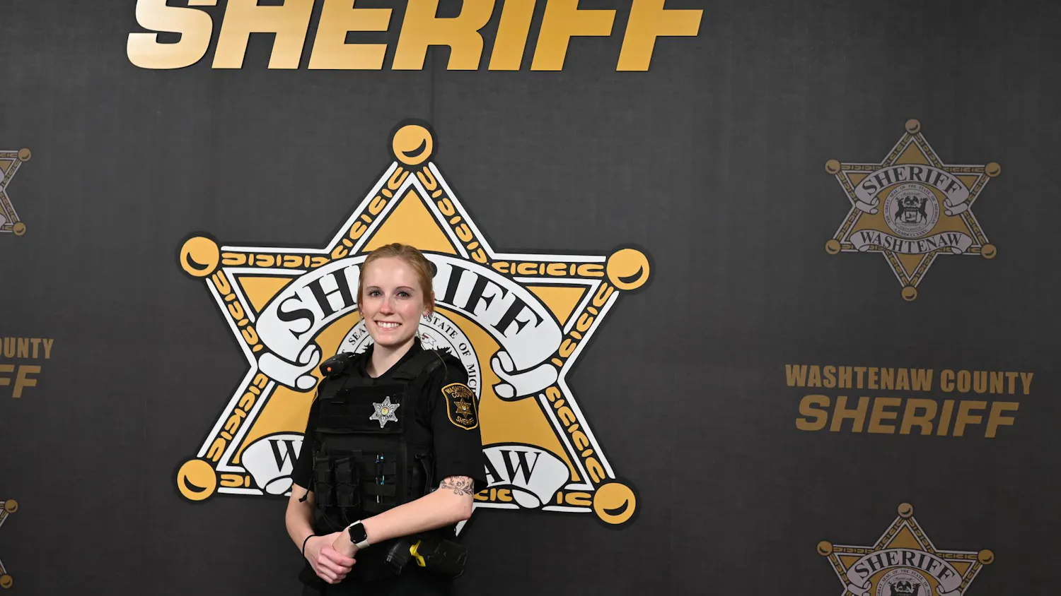 Jessica VanBynen stands in uniform in front of a backdrop with the Washtenaw County Sheriff's Office logo.