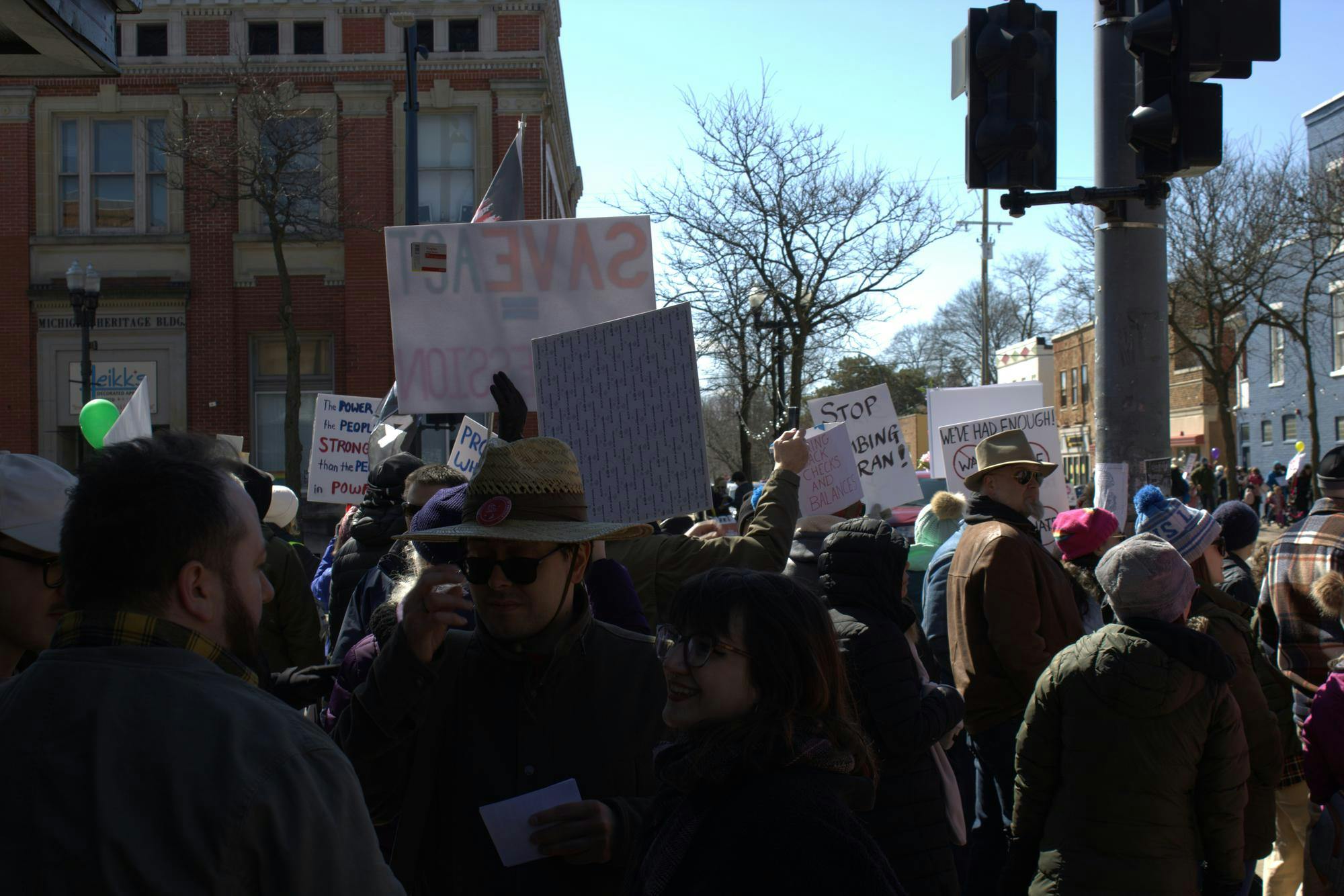 Dozens of people stand on a sidewalk beside a road in downtown Ypsilanti, some holding signs up, facing cars driving past. 