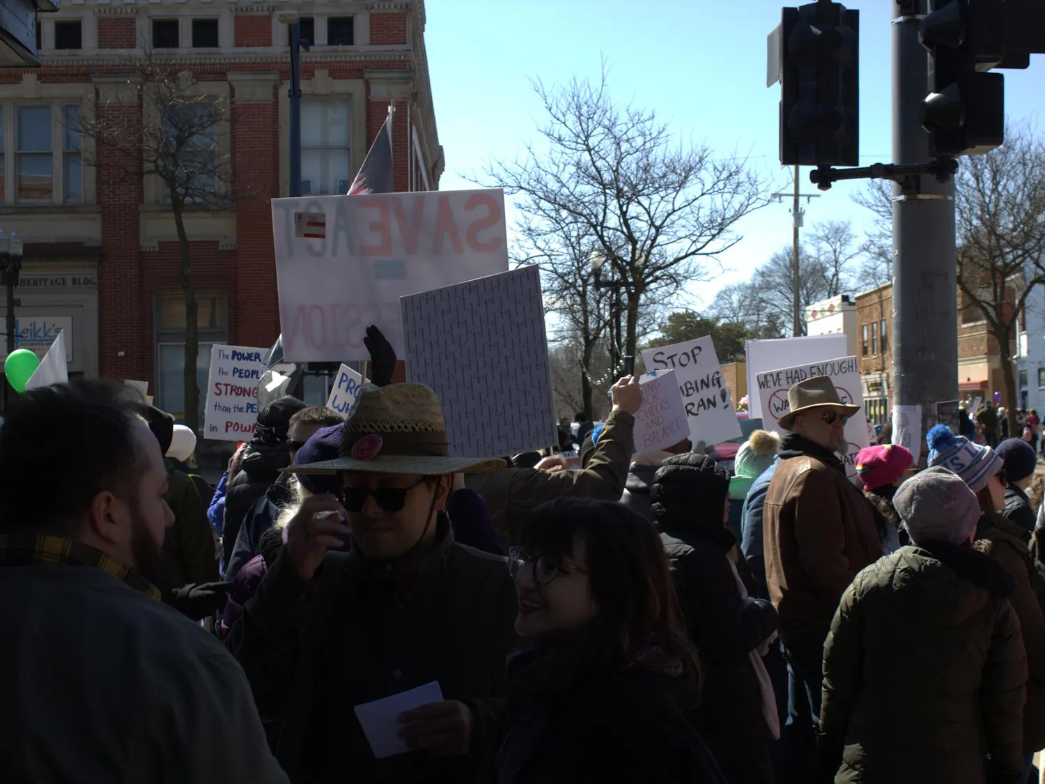Dozens of people stand on a sidewalk beside a road in downtown Ypsilanti, some holding signs up, facing cars driving past.