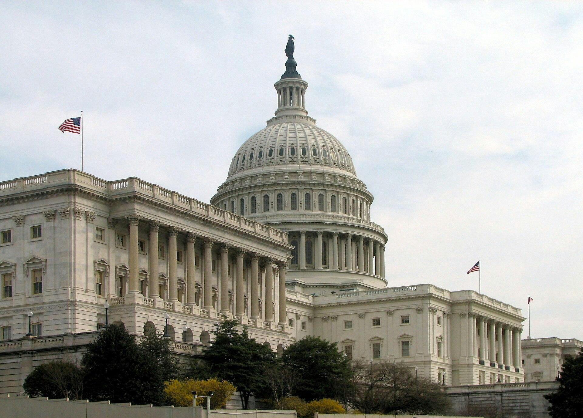 The U.S. Capitol building features a dome, columns and other aspects of Greek and Roman architecture, all in a white color; American flags fly from the building.