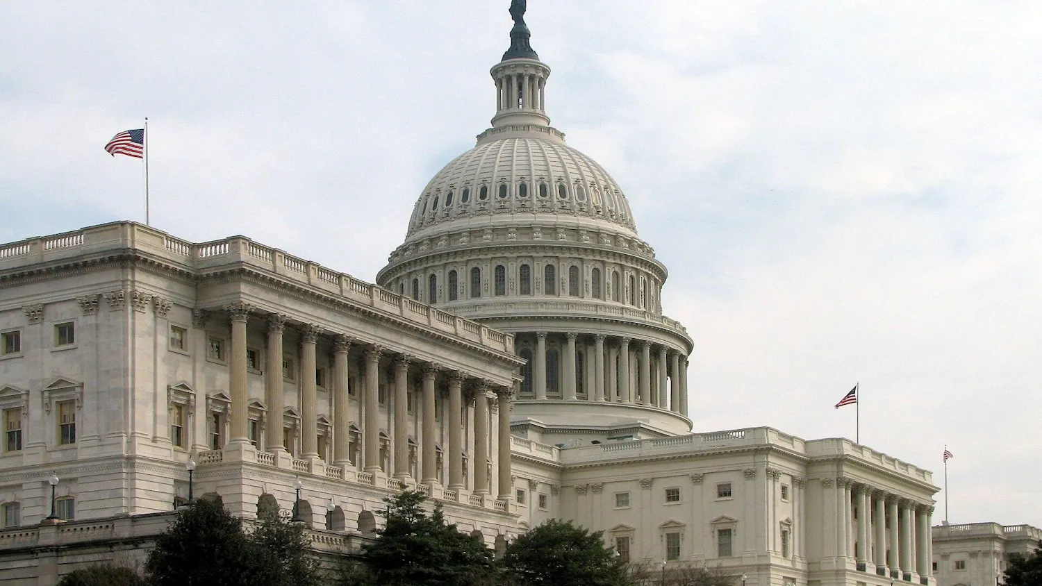 The U.S. Capitol building features a dome, columns and other aspects of Greek and Roman architecture, all in a white color; American flags fly from the building.