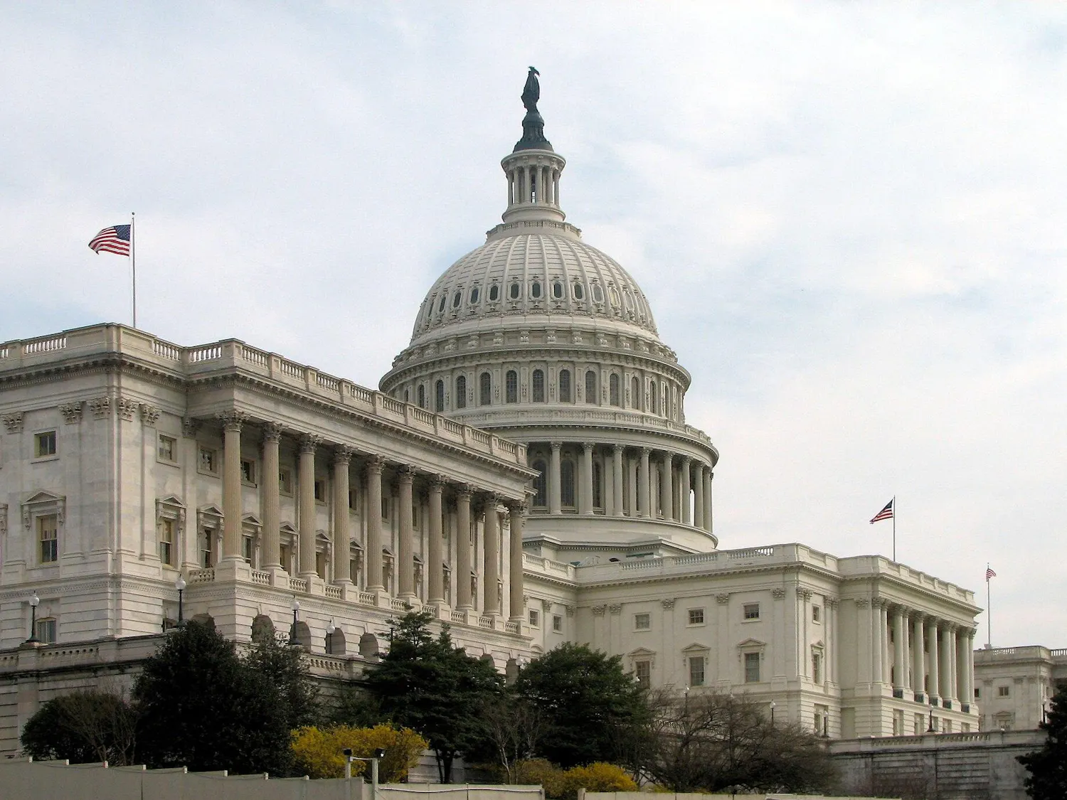 The U.S. Capitol building features a dome, columns and other aspects of Greek and Roman architecture, all in a white color; American flags fly from the building.