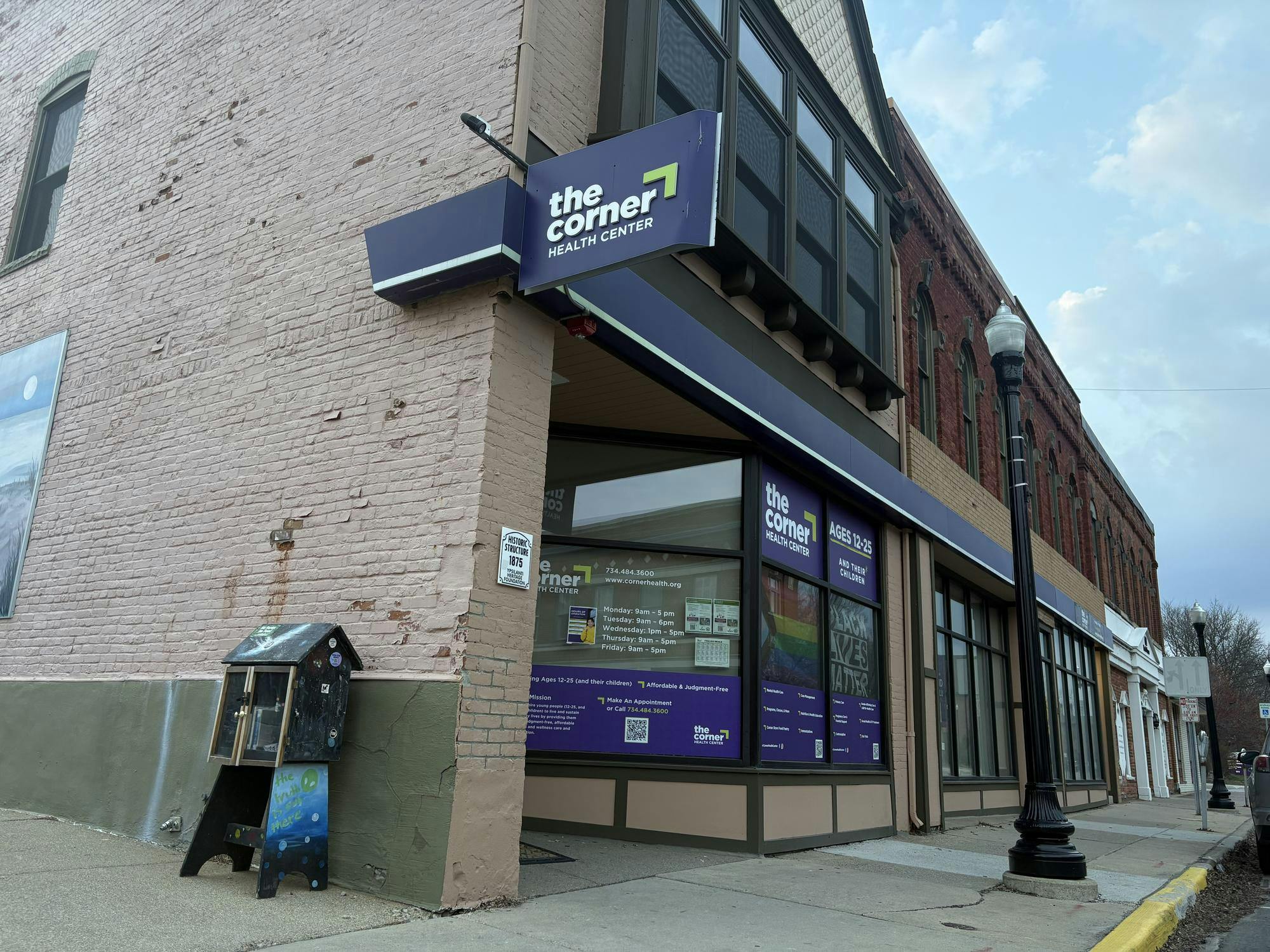 Storefront on the street corner of a block of businesses. The side of the building is made of beige bricks and features large windows in the front. The sign above the door reads, "The Corner Health Center."