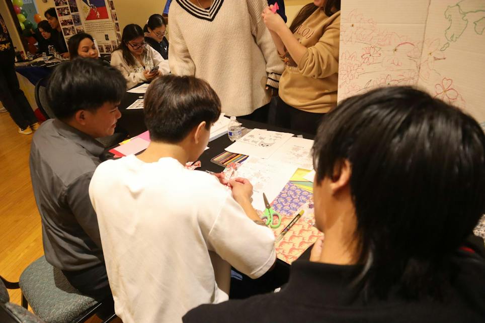Students are seen from over the shoulder at a table covered in colorful and patterned paper and the students are cutting and folding the paper.