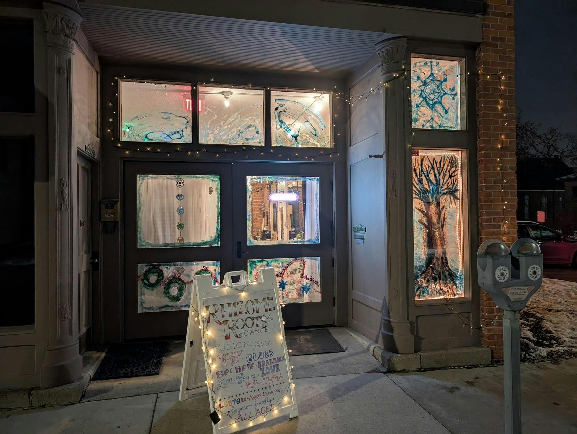 The storefront of a building decorated for the winter holidays. Snowflakes, green wreaths and other winter-themed items are painted on the glass doors and windows of the building. In front of the building is a white sign that says "Rhizome Roots" in gold and red letters. It is night, and the lights in the building are on.