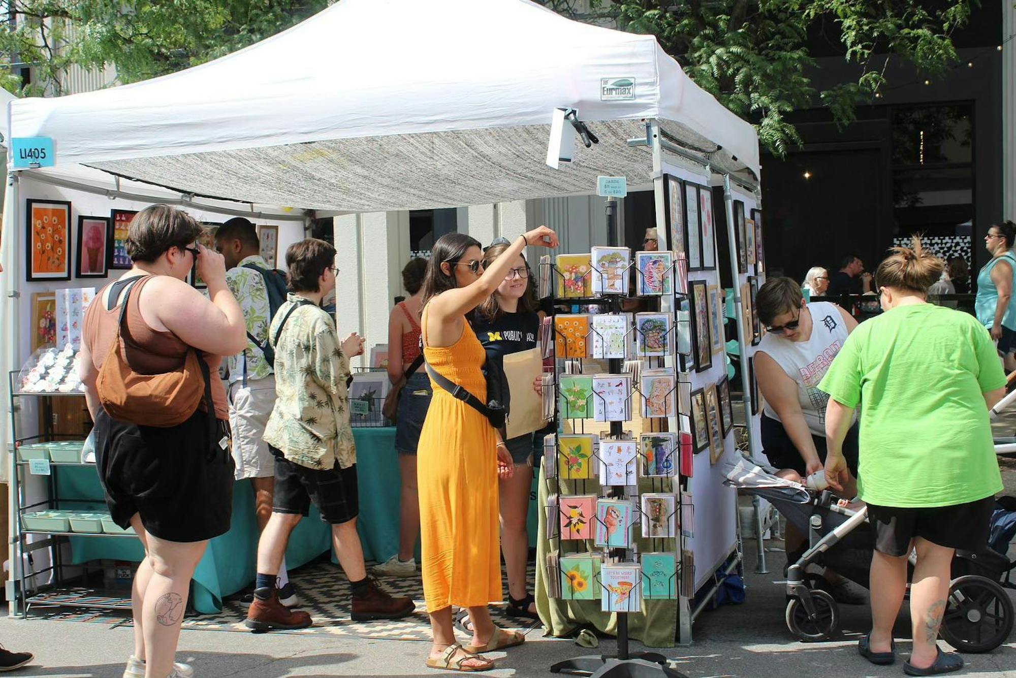 Art prints and other works are set up under a white tent outside. People stand in and around the tent looking through the artwork and picking up items. It is a sunny day and people are dressed for summer.