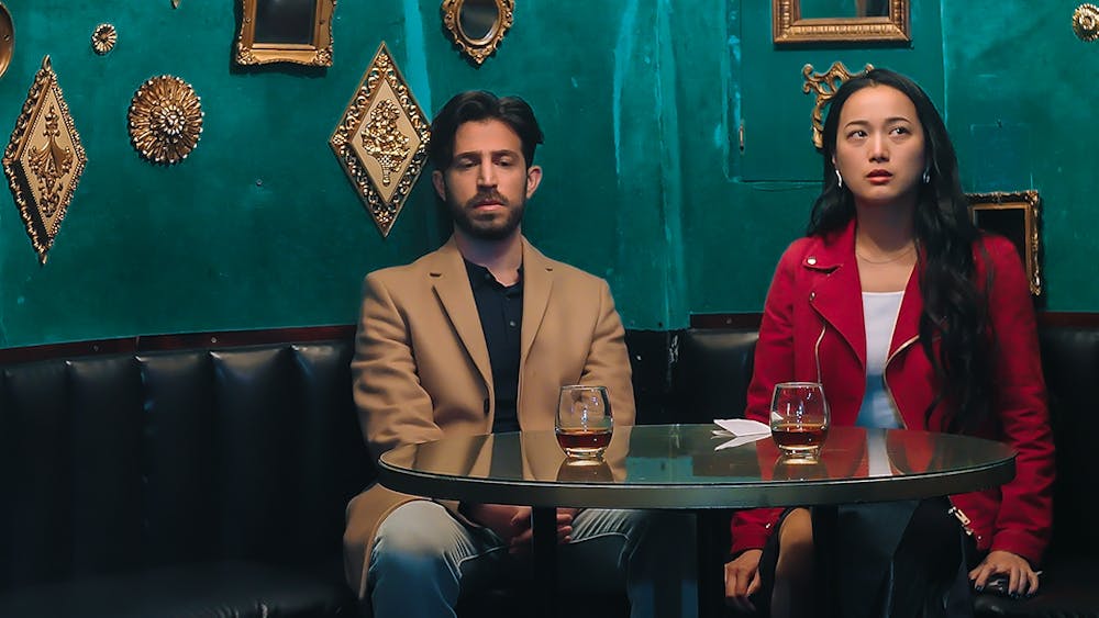 A man and woman seated at a booth. Behind them is a blue wall with golden colored frames scattered around. The man and the woman sit awkwardly, not looking at one another. The man wears a tan suit, and the woman wears a pink jacket over a white shirt.