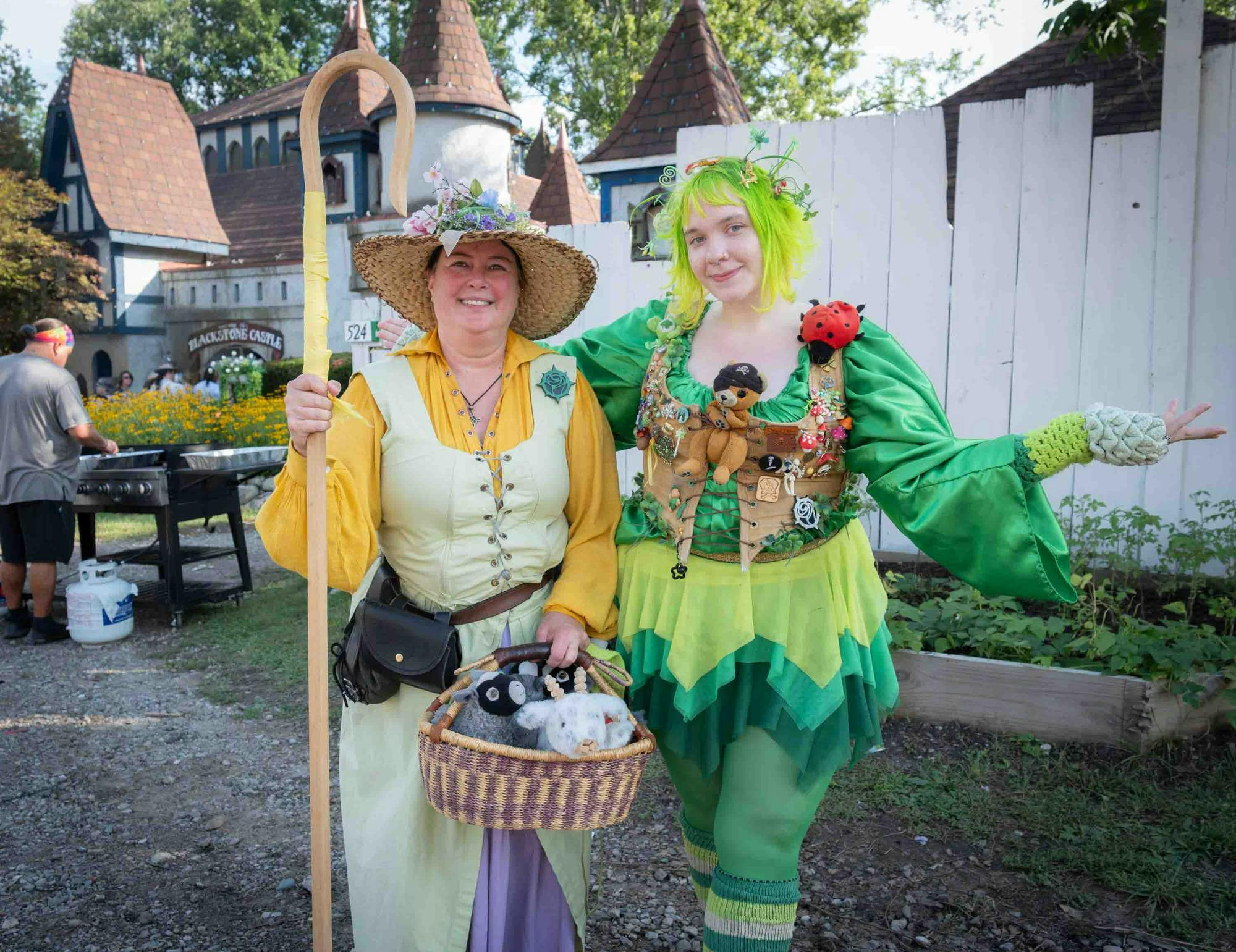 Two people dressed in renaissance attire pose on festival grounds, along with accessories including a walking stick and small stuffed animals.