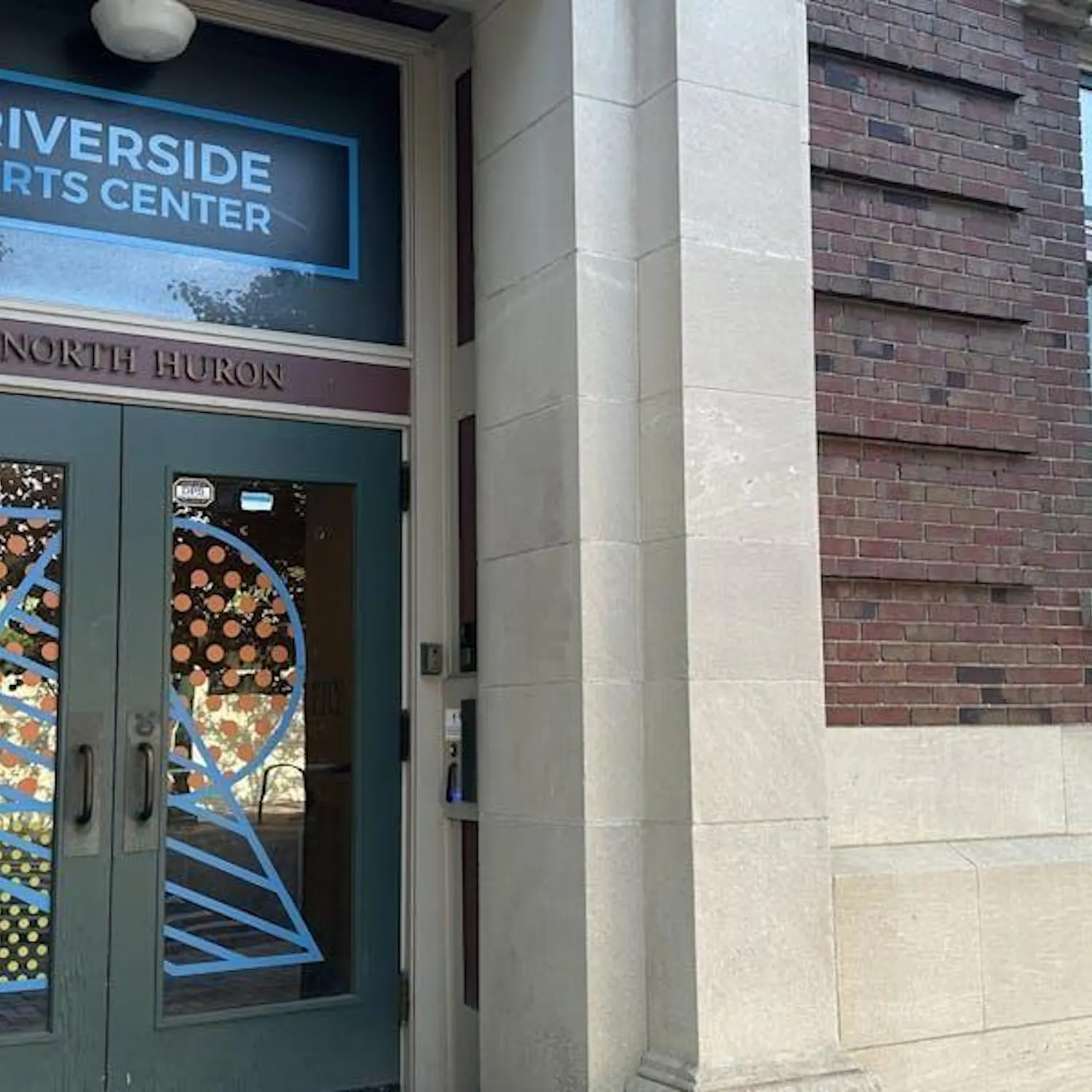 Doorway to a gray cement and red brick historic building. The glass double-door entrance has the letter "R" painted on the front in colorful patterns.
