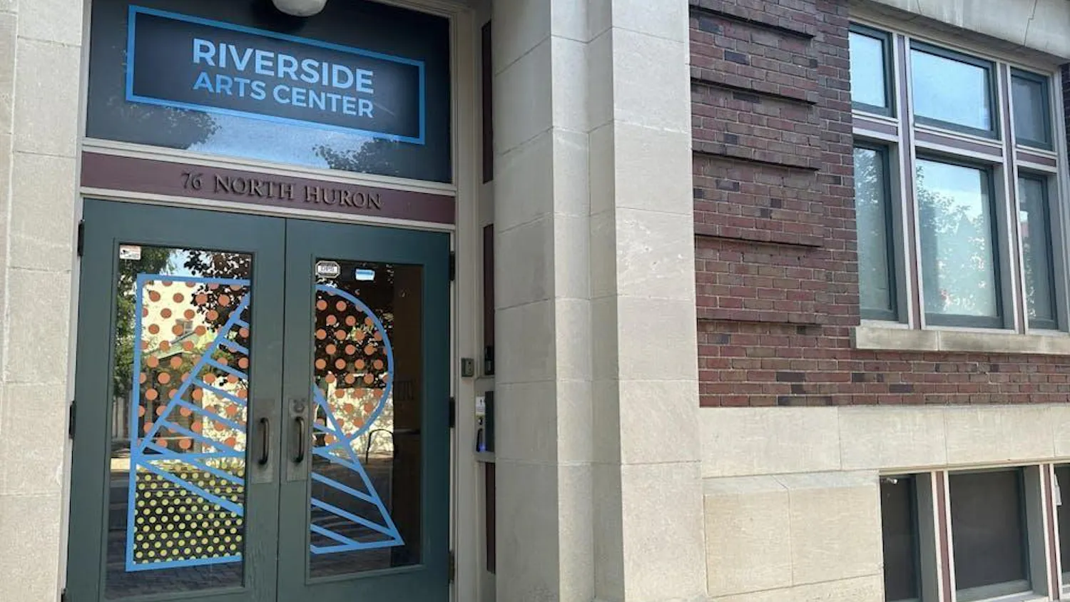 Doorway to a gray cement and red brick historic building. The glass double-door entrance has the letter "R" painted on the front in colorful patterns.