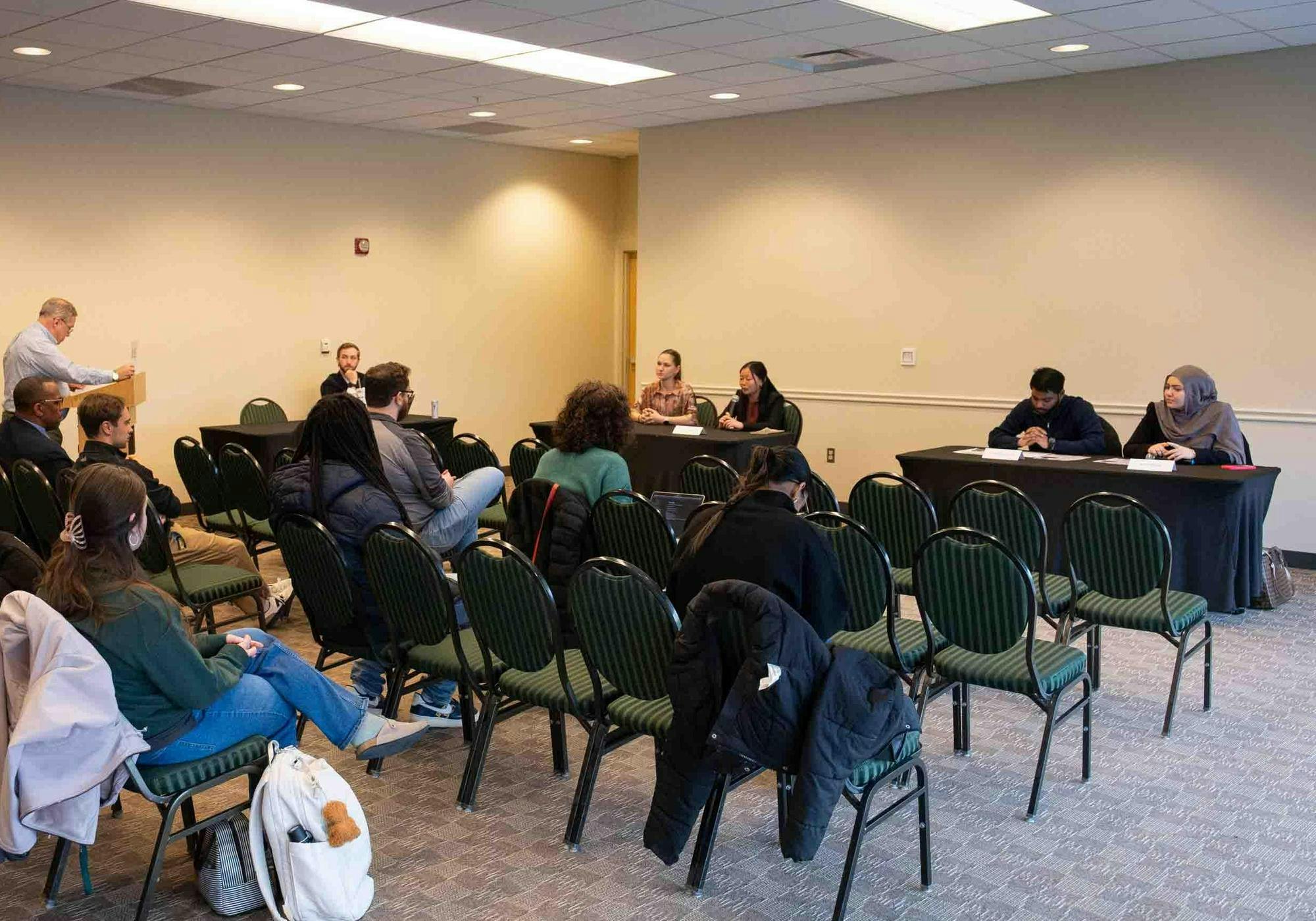 At two tables, candidates from each presidential ticket sit and answer questions. Attendees watch from rows of chairs.