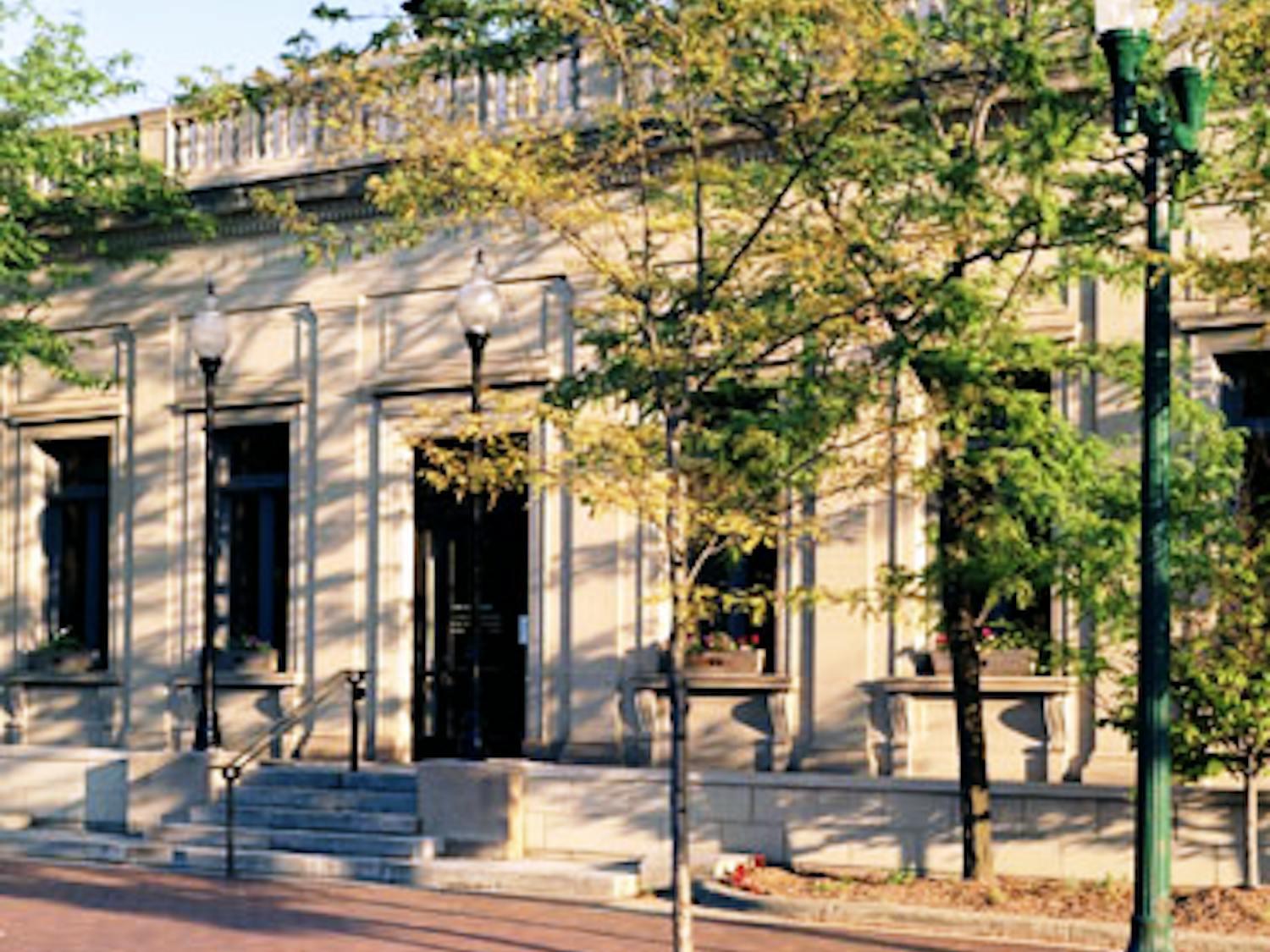 The front of a tan-colored building with many dark windows and several steps leading up to the door. It is a sunny day, and the trees in the foreground are casting shadows onto the building.