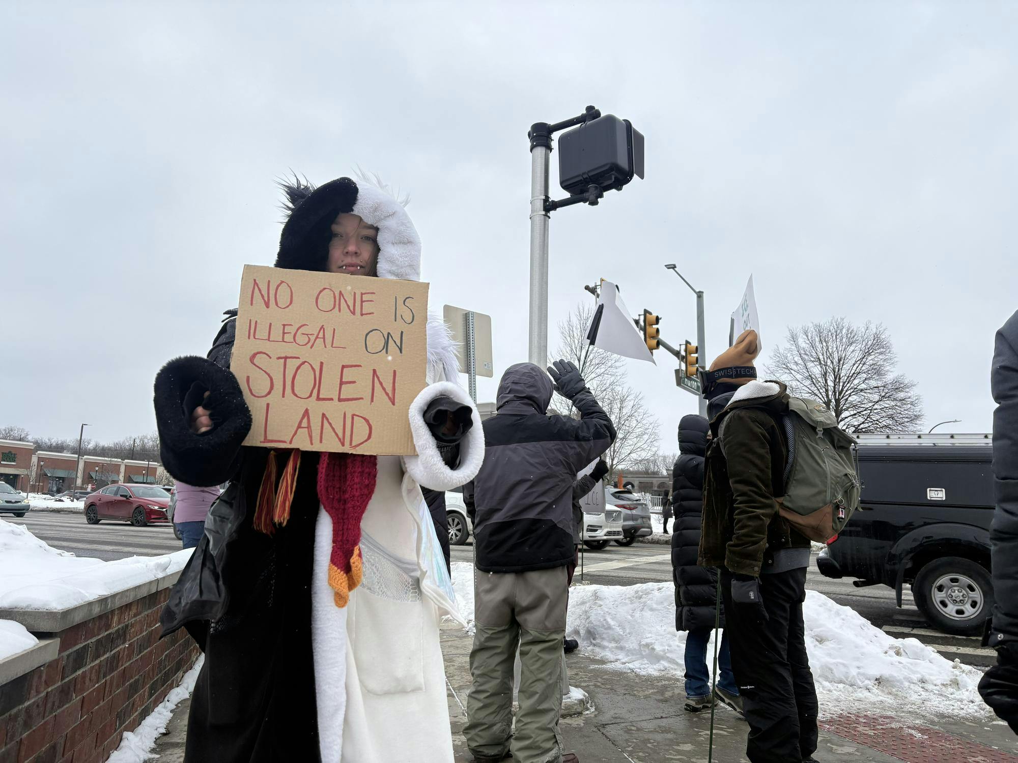 Hannah Blackburn wears a black and white furry coat and holds a sign that says "No one is illegal on stolen land." Behind them, other protesters stand on the sidewalk along the intersection while cars drive by.