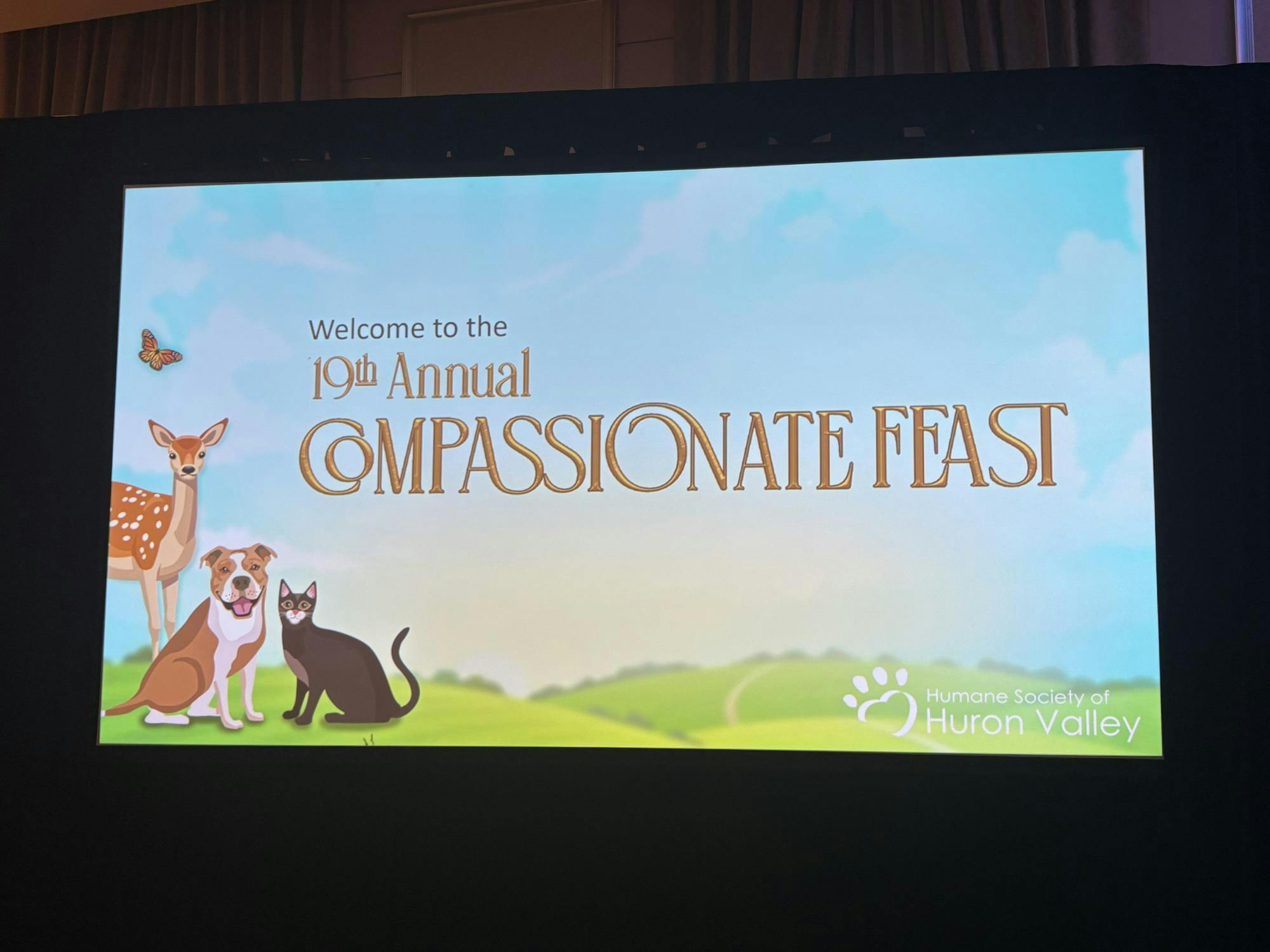 A screen displays and image of green hills below a blue sky. A deer, dog, cat, and butterfly sit at the bottom left corner. Across the screen in gold cursive it says "Welcome to the 19th annual Compassionate Feast" and in the bottom right corner there is white text that says "Humane Society of Huron Valley" with their paw logo next to it. 