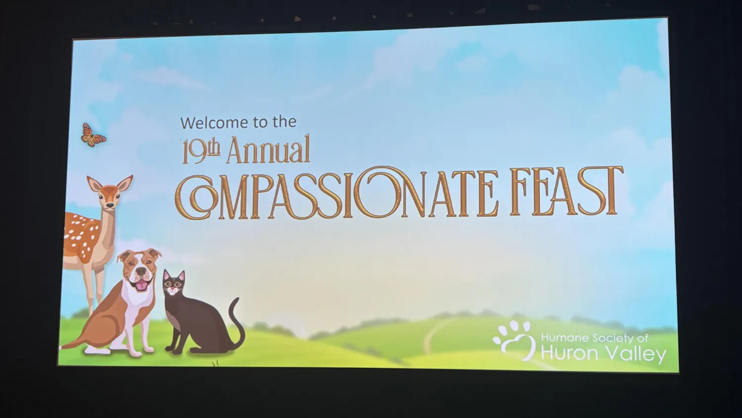 A screen displays and image of green hills below a blue sky. A deer, dog, cat, and butterfly sit at the bottom left corner. Across the screen in gold cursive it says "Welcome to the 19th annual Compassionate Feast" and in the bottom right corner there is white text that says "Humane Society of Huron Valley" with their paw logo next to it.