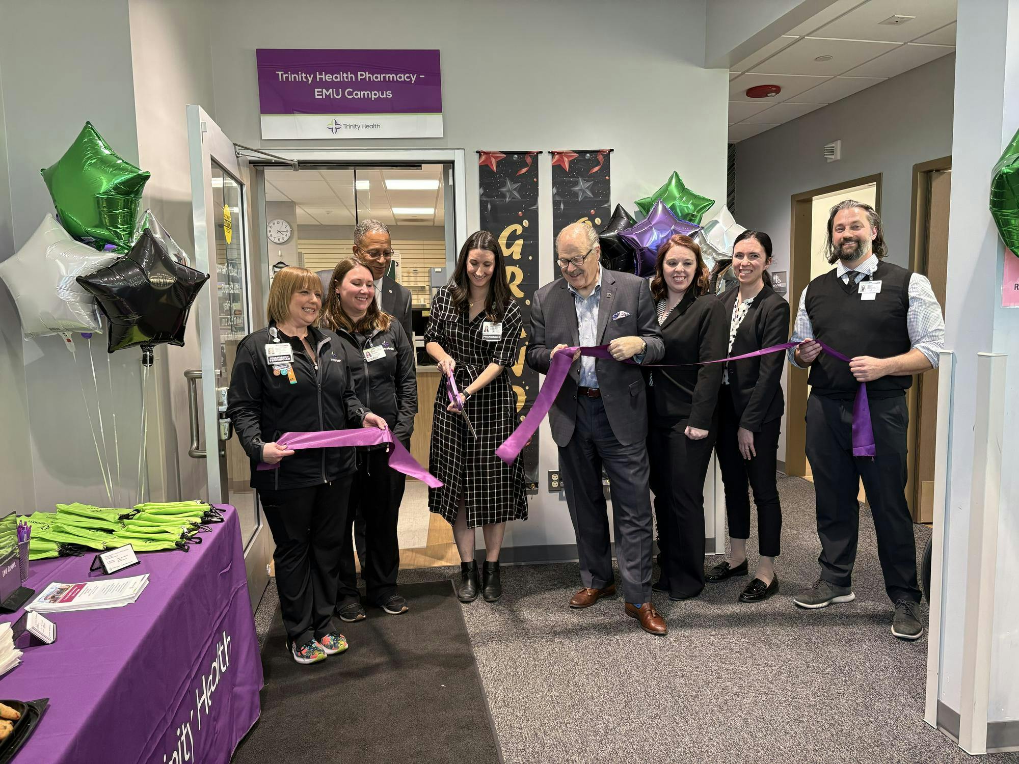 EMU President James Smith and pharmacy staff hold a purple ribbon while one person cuts the ribbon with a large pair of purple scissors.