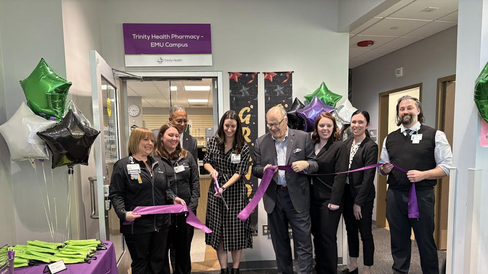 EMU President James Smith and pharmacy staff hold a purple ribbon while one person cuts the ribbon with a large pair of purple scissors.