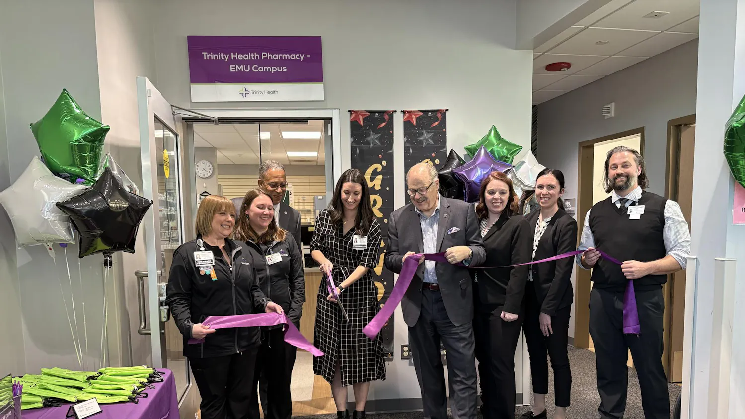 EMU President James Smith and pharmacy staff hold a purple ribbon while one person cuts the ribbon with a large pair of purple scissors.