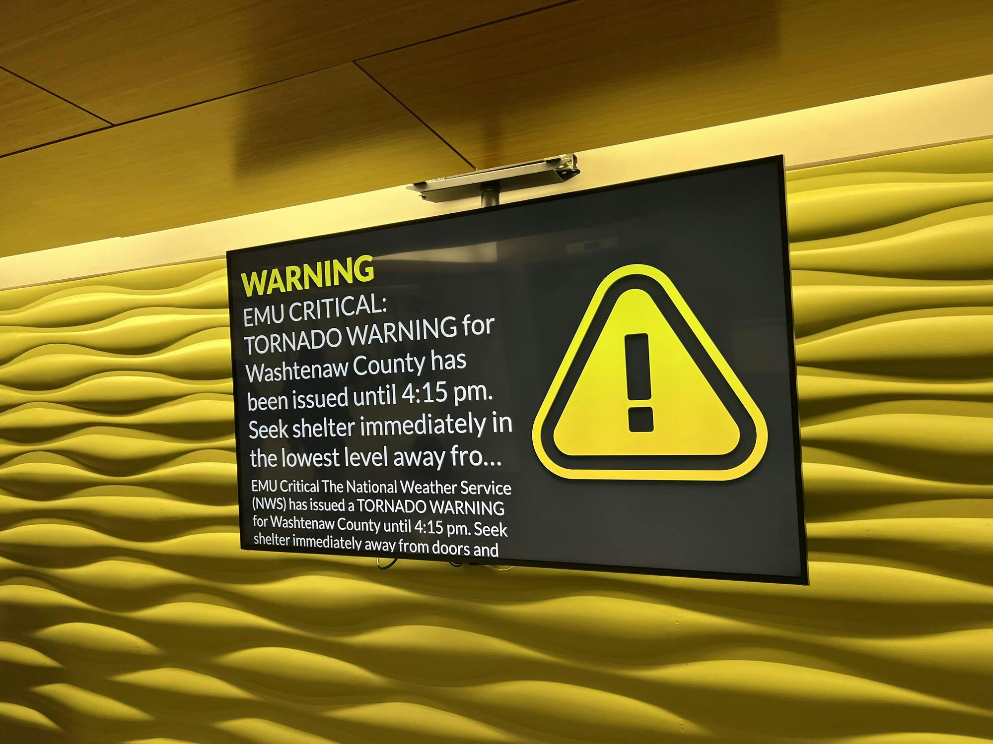 A TV screen shows an alert that says, "EMU Critical: Tornado Warning for Washtenaw County has been issued until 4:15 pm. Seek shelter immediately on the lowest level away from doors and windows." Next to the text there is a large yellow triangle with a black exclamation point inside.