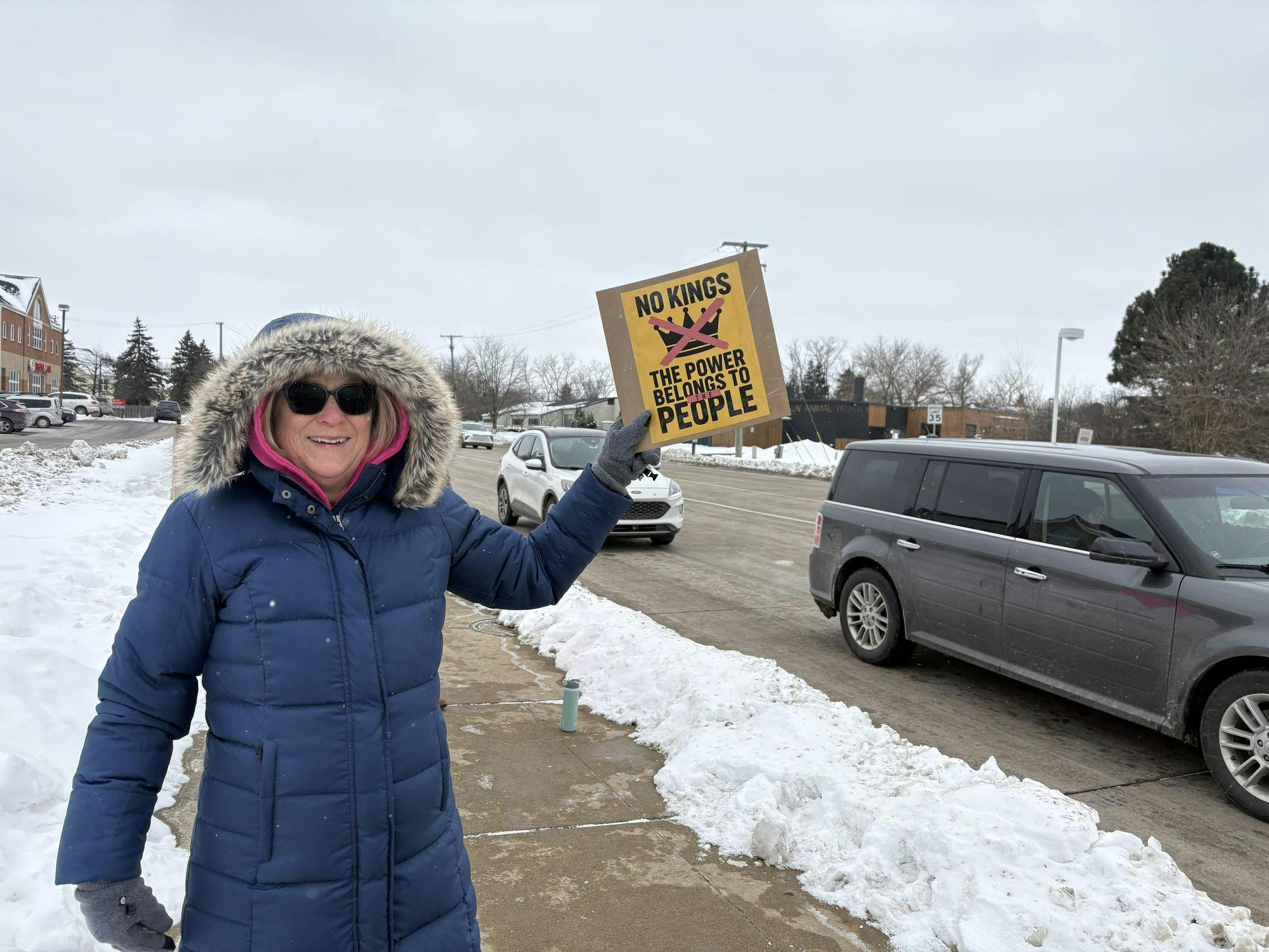 Cheryl Brooks is wearing a blue winter coat and smiling while holding a yellow sign that says "No Kings, the power belongs to people" with a picture of a crown crossed out. In the background, cars drive by in the street behind her. 