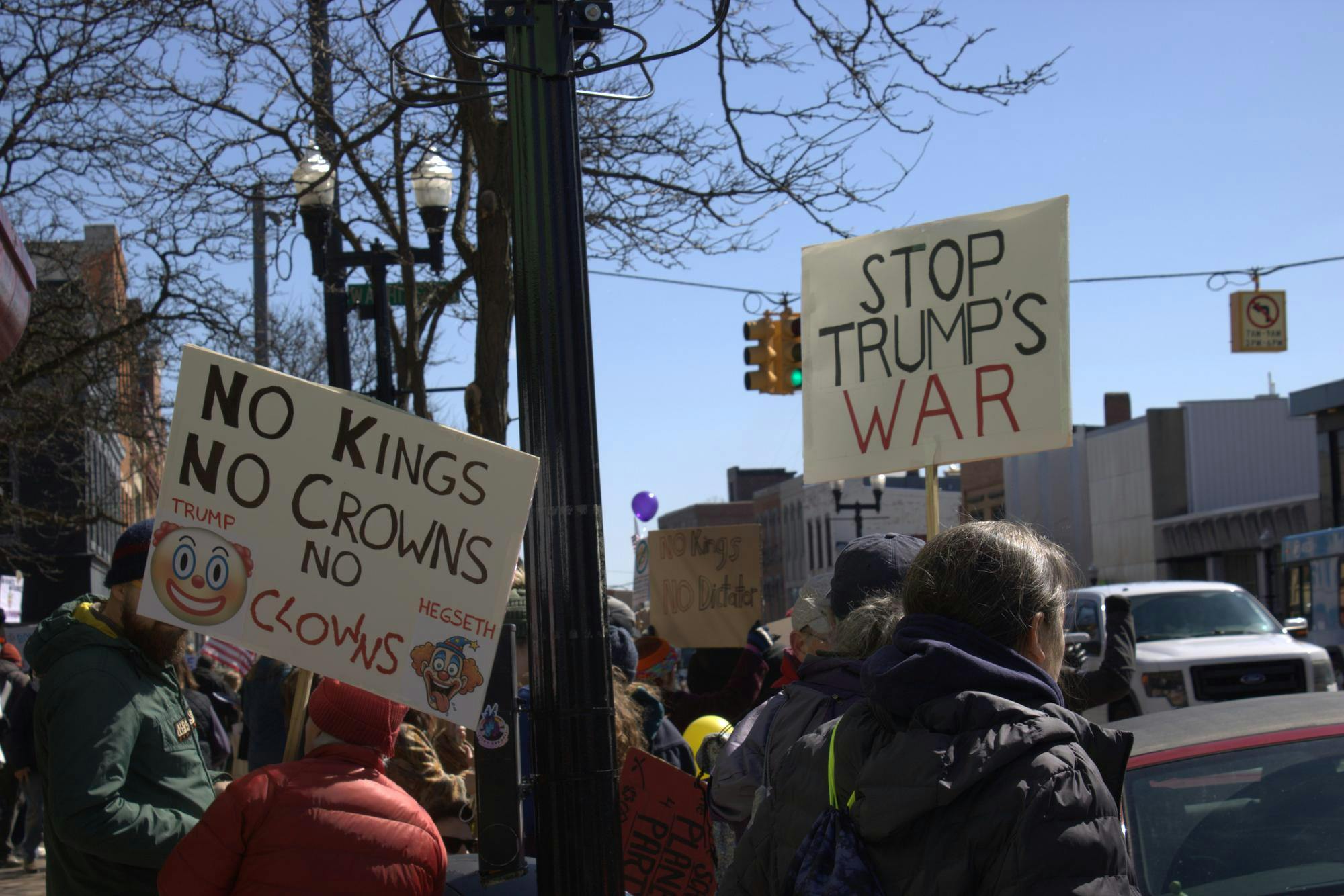 Protesters hold up two signs, one reading "No kings, no crowns," with printed clown emojis and images, and the other reading "stop trump's war." They stand among a crowd of people on a sidewalk beside a road. 