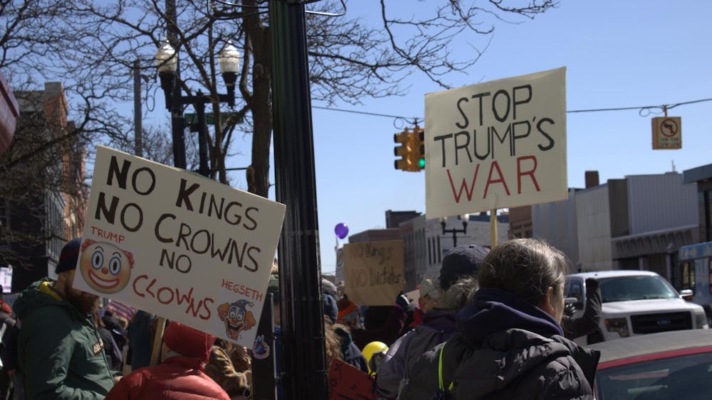 Protesters hold up two signs, one reading "No kings, no crowns," with printed clown emojis and images, and the other reading "stop trump's war." They stand among a crowd of people on a sidewalk beside a road.