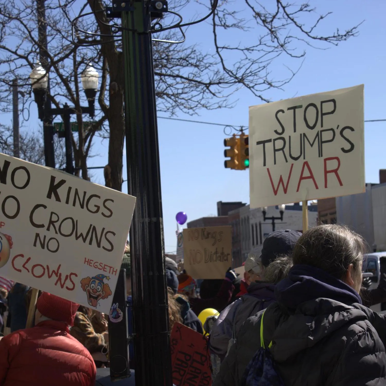 Protesters hold up two signs, one reading "No kings, no crowns," with printed clown emojis and images, and the other reading "stop trump's war." They stand among a crowd of people on a sidewalk beside a road.