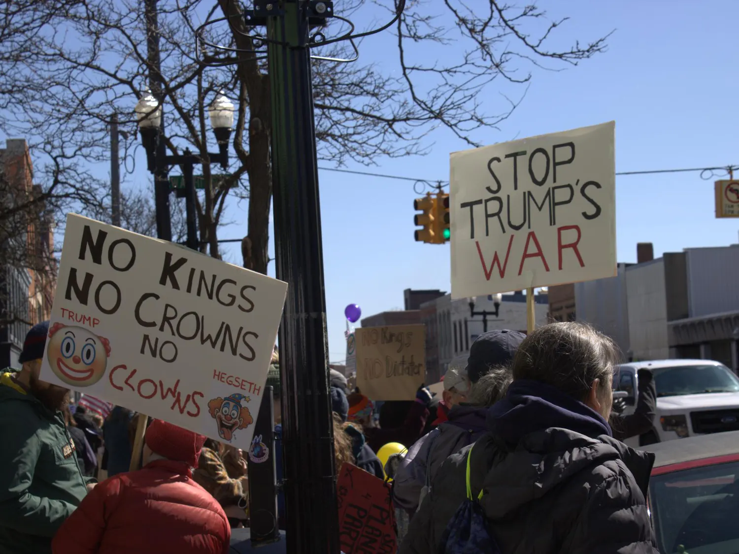 Protesters hold up two signs, one reading "No kings, no crowns," with printed clown emojis and images, and the other reading "stop trump's war." They stand among a crowd of people on a sidewalk beside a road.