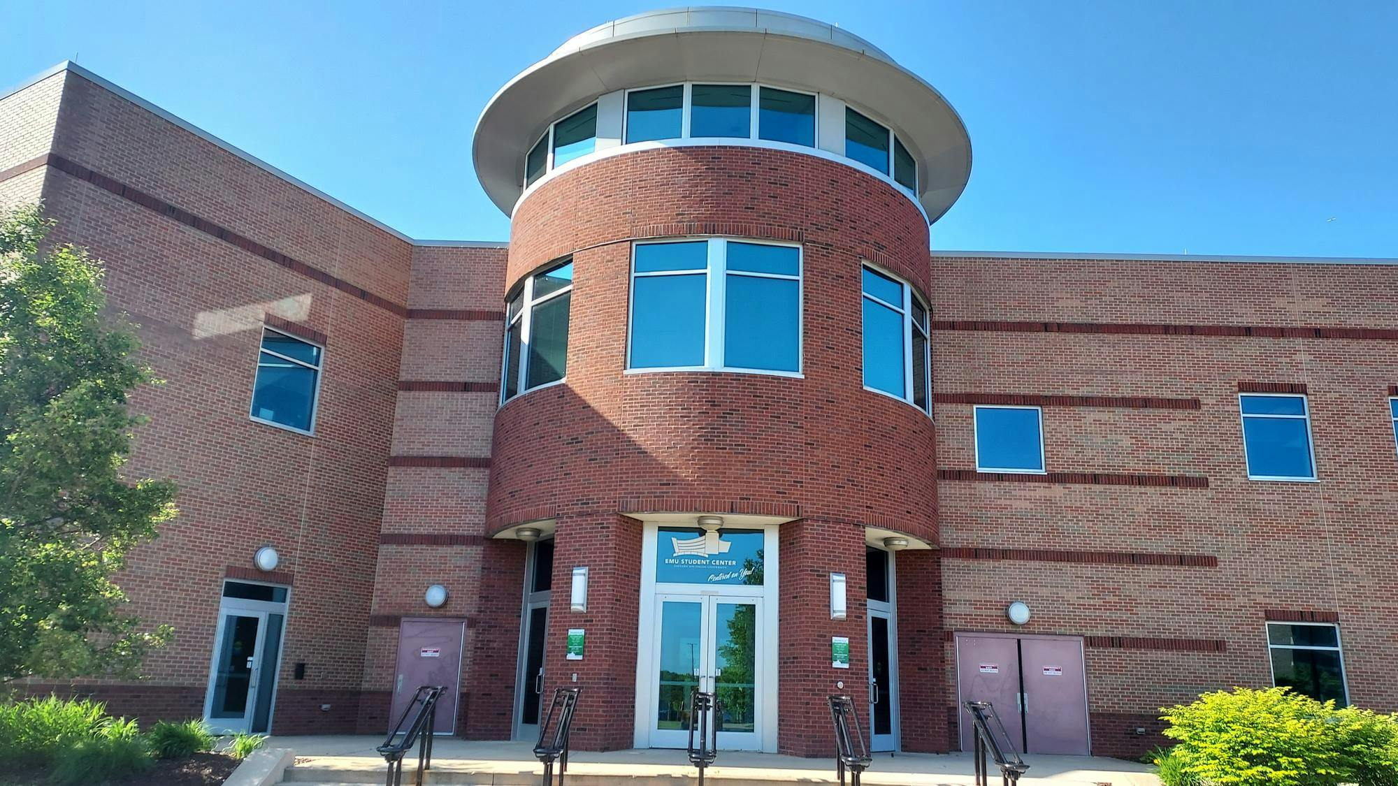 The entrance of the EMU student center has a few stairs to step on and three front doors to enter. The building is made of red brick walls and glass windows. 