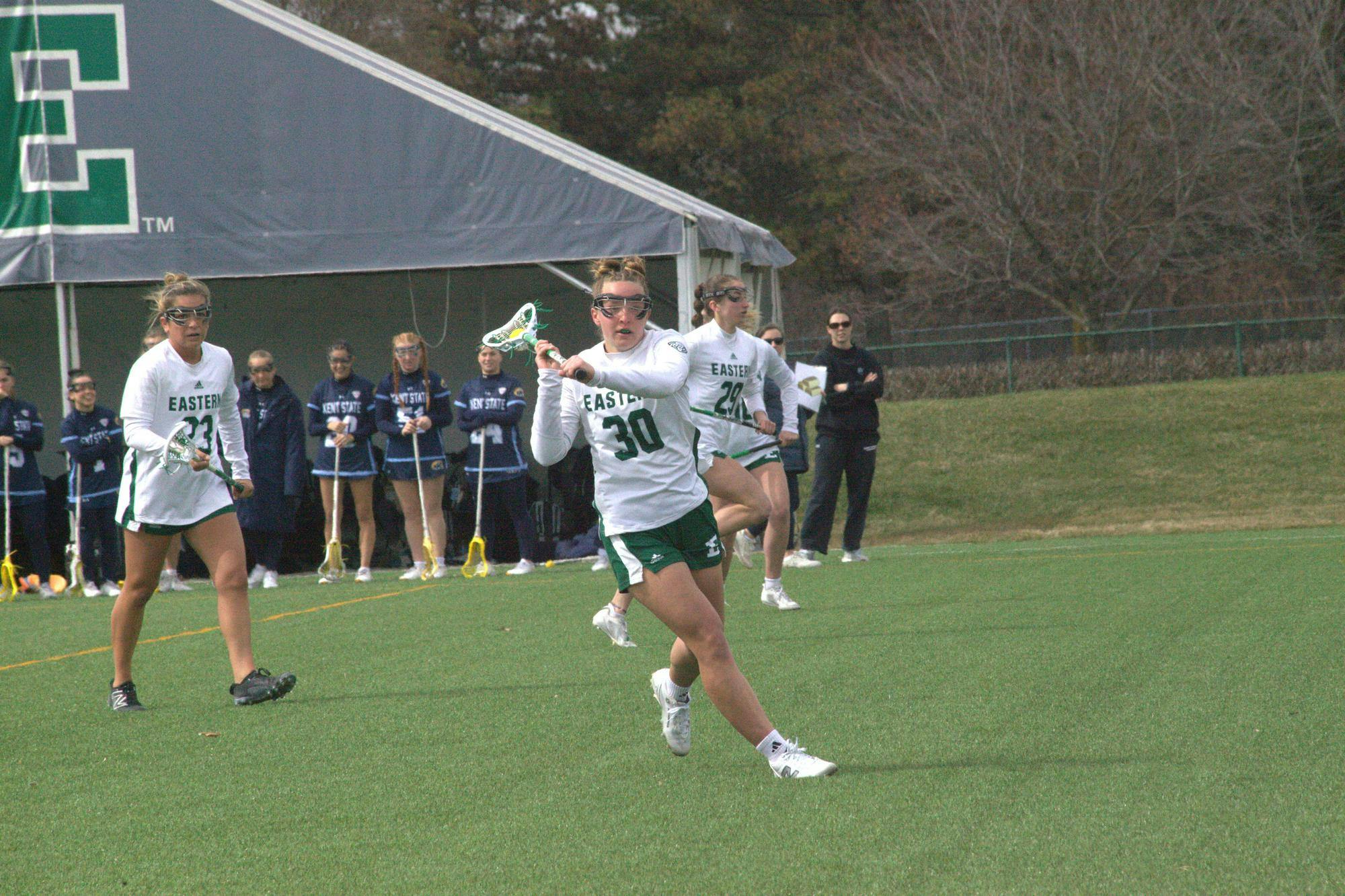 Women's lacrosse player prepares to pass the ball to teammate who is off-screen on outdoor lacrosse field while members of both teams watch in the background.