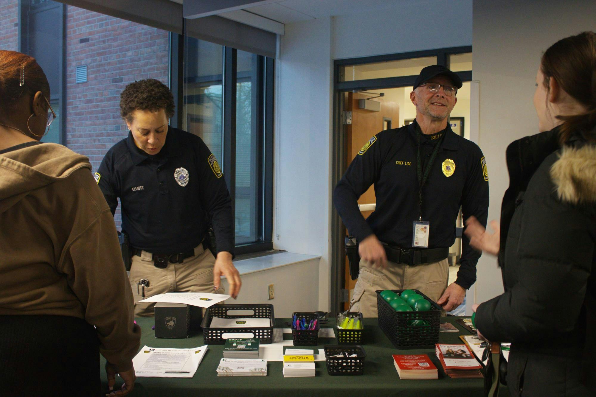 Two campus police officers in uniform talk to two Eastern Michigan students while standing behind an information table.