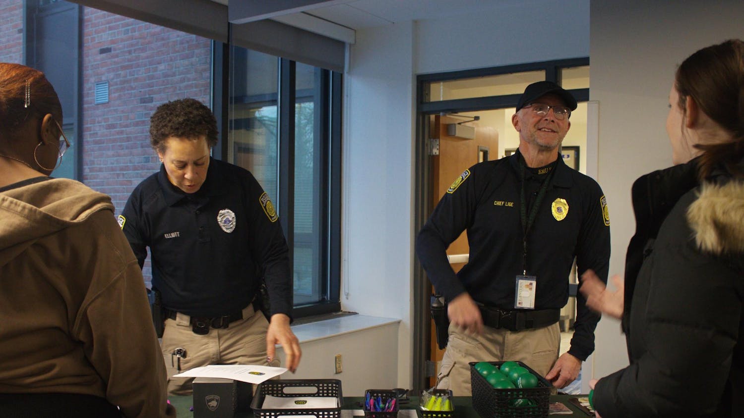 Two campus police officers in uniform talk to two Eastern Michigan students while standing behind an information table.