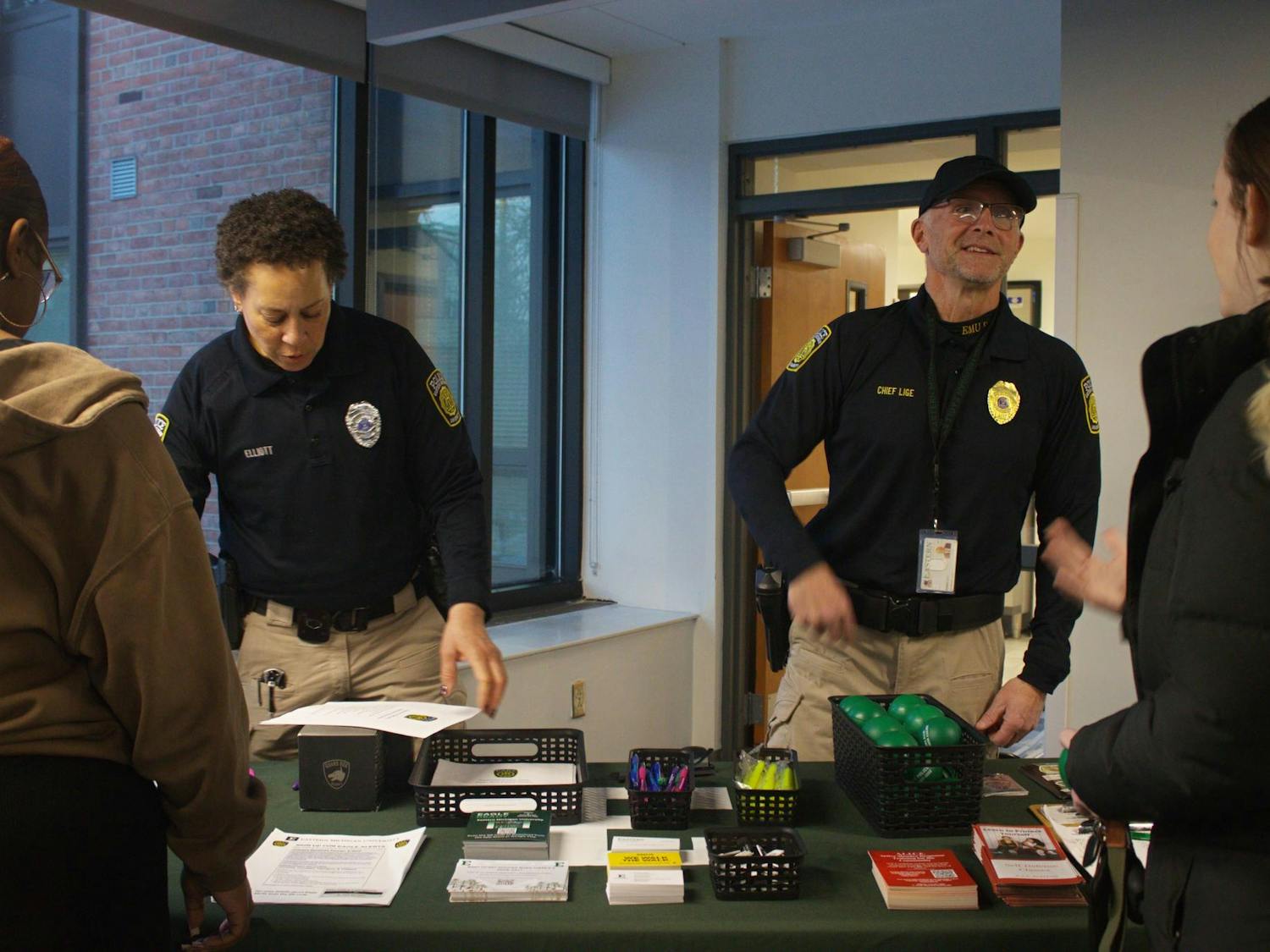 Two campus police officers in uniform talk to two Eastern Michigan students while standing behind an information table.