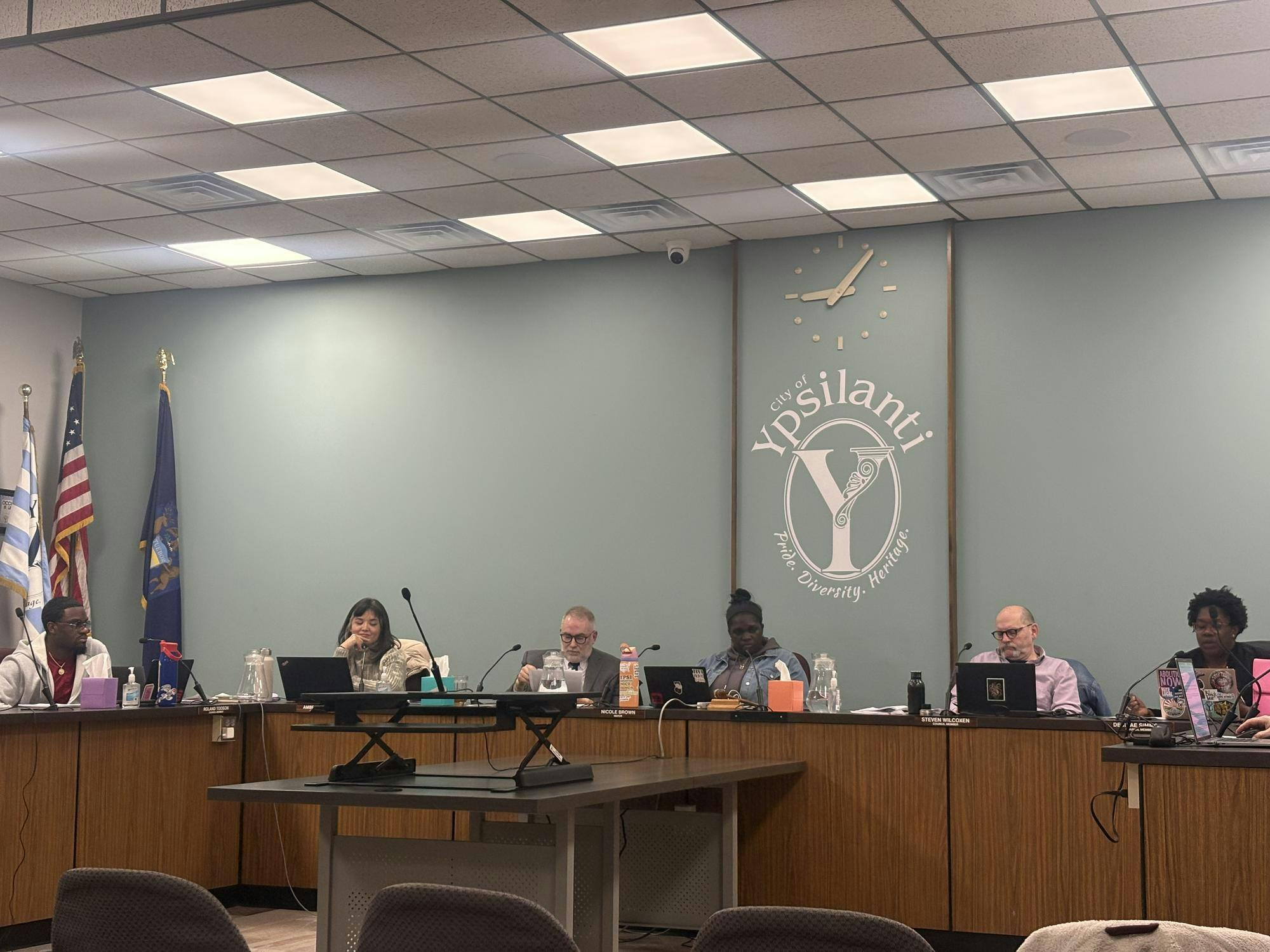 6 council members sit behind a large, wooden, U-shaped desk. The "Y" shaped logo of Ypsilanti is painted on the green wall behind them.