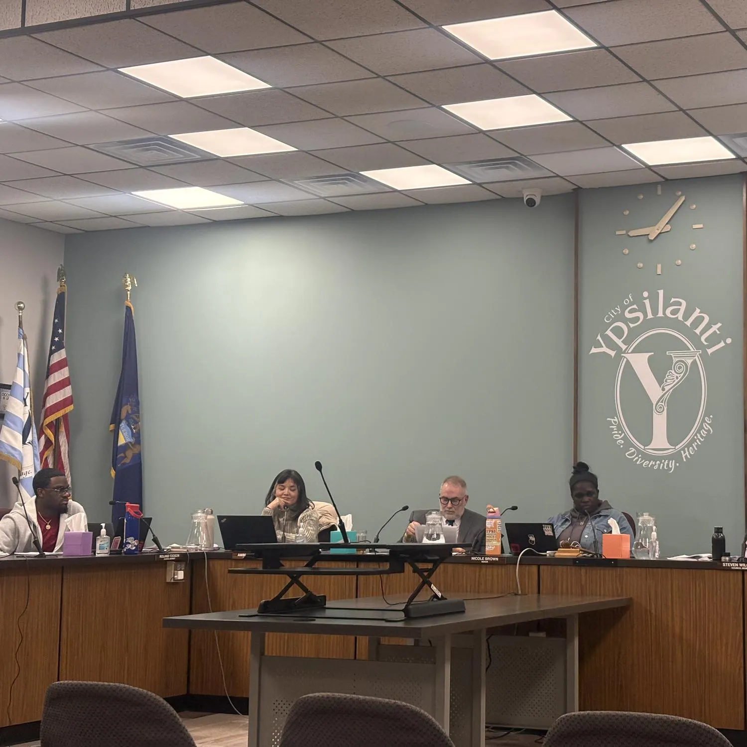 6 council members sit behind a large, wooden, U-shaped desk. The "Y" shaped logo of Ypsilanti is painted on the green wall behind them.