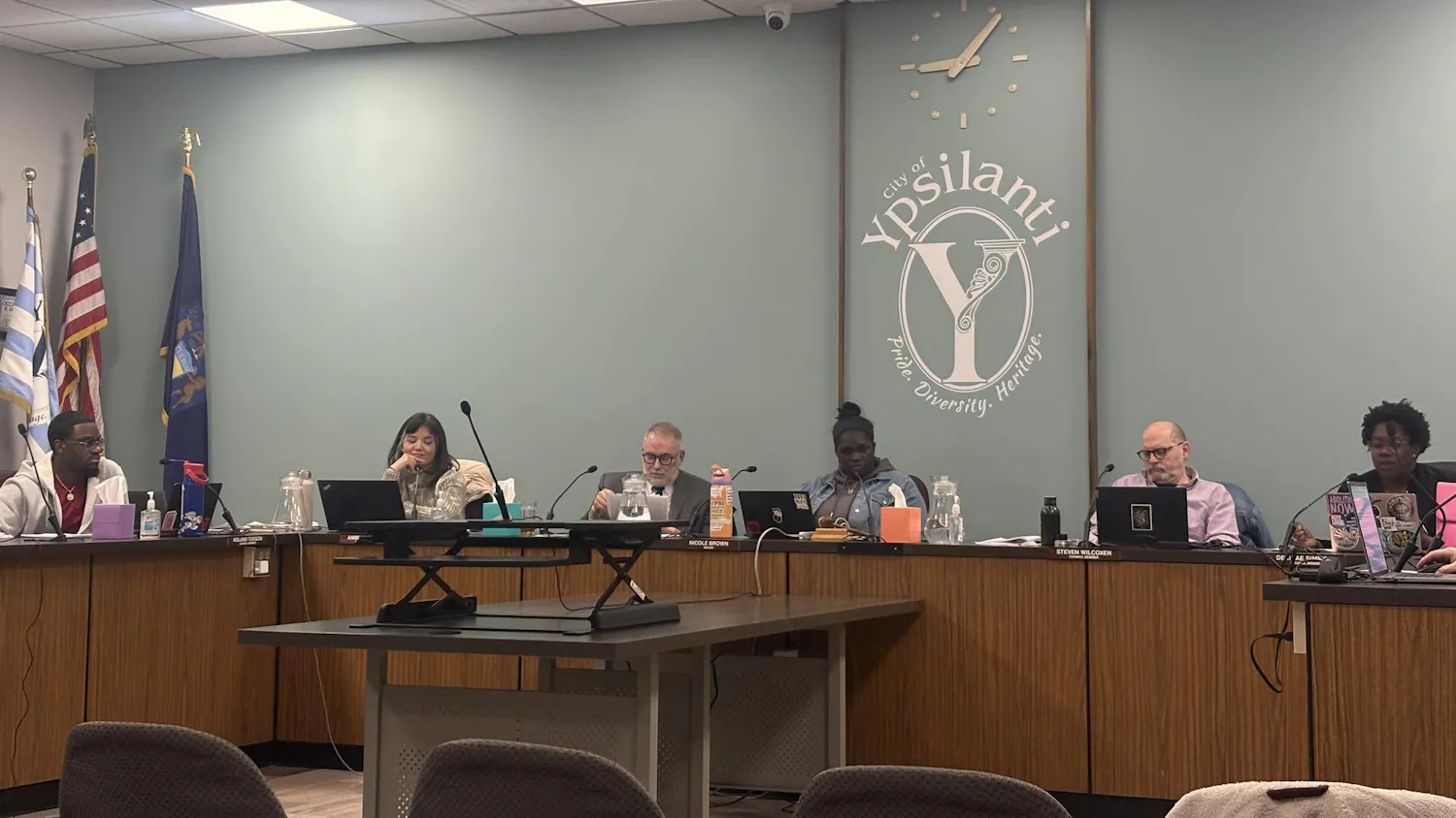 6 council members sit behind a large, wooden, U-shaped desk. The "Y" shaped logo of Ypsilanti is painted on the green wall behind them.