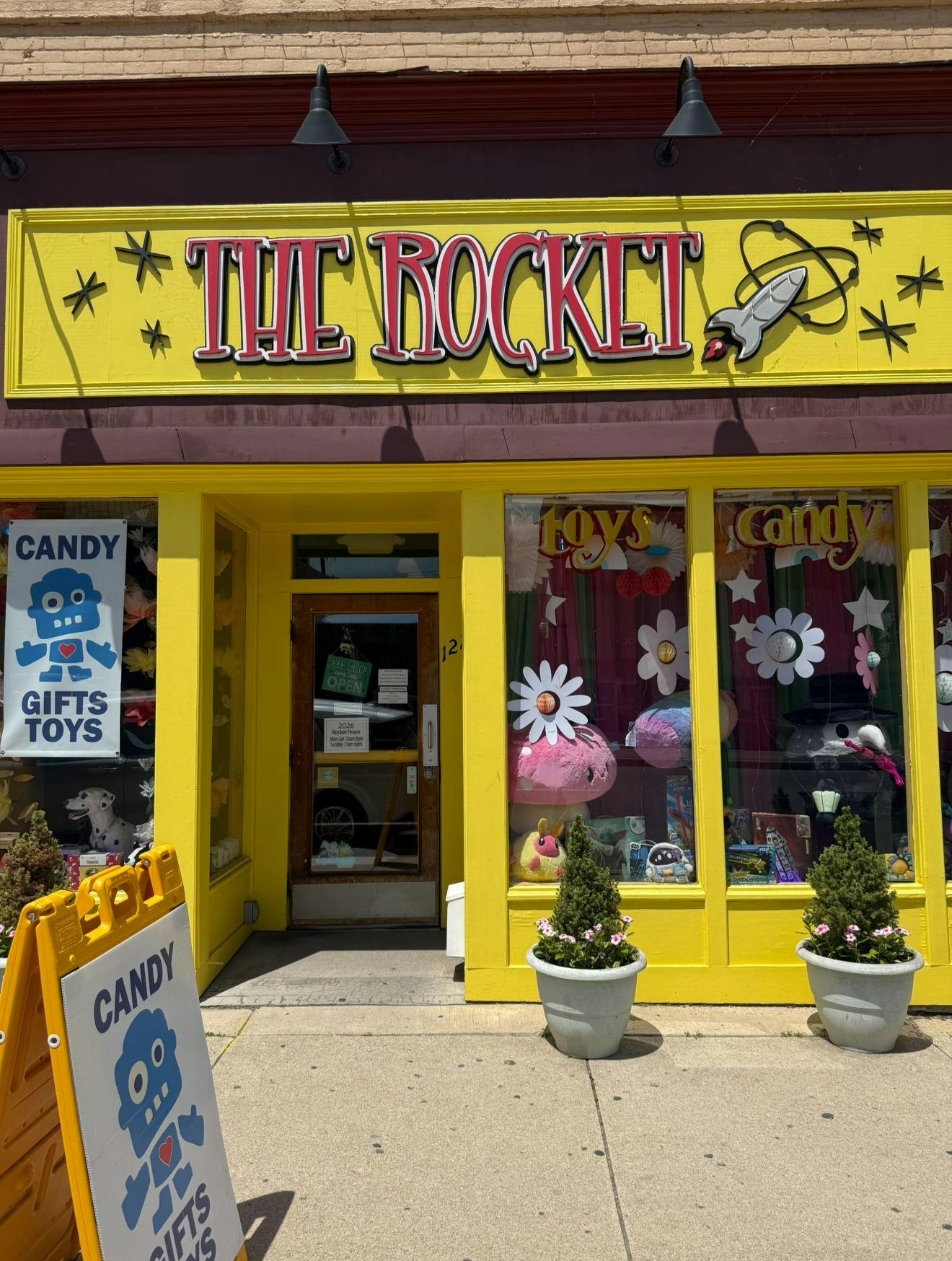 The Rocket's yellow storefront has the words "The Rocket" and is covered with daisies on the window, large plush characters on display inside the window, and a sign that says, "Candy, Gifts, Toys."