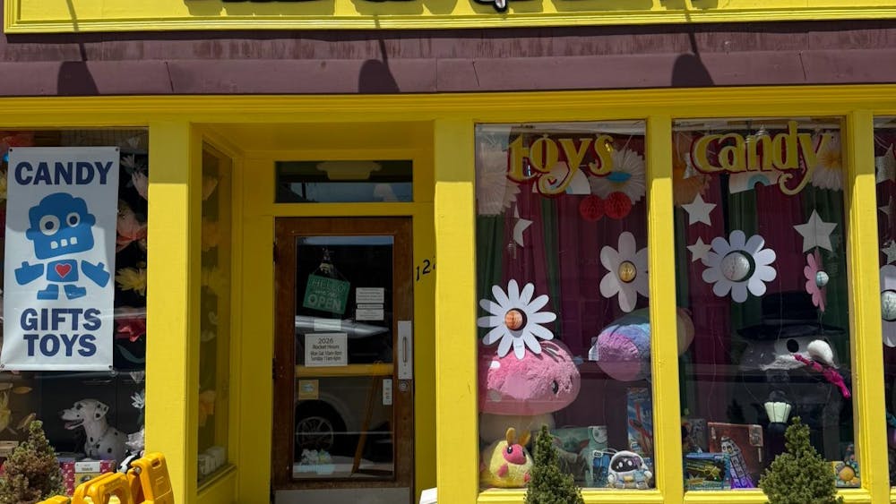 The Rocket's yellow storefront has the words "The Rocket" and is covered with daisies on the window, large plush characters on display inside the window, and a sign that says, "Candy, Gifts, Toys."