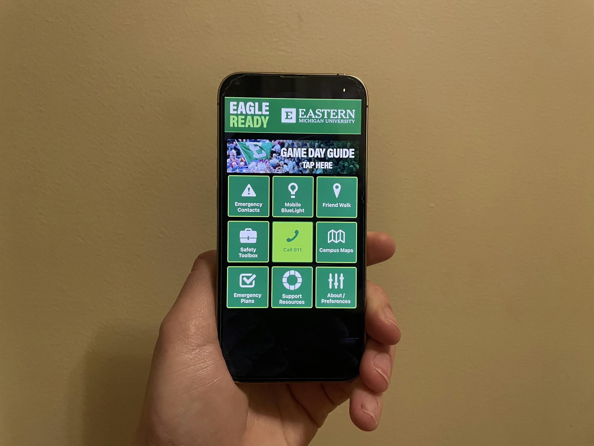 A hand holding a phone that is displaying a black and green app. The home page has buttons for various emergency services and campus resources.