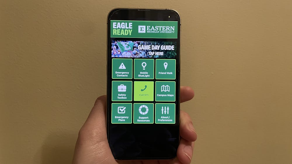 A hand holding a phone that is displaying a black and green app. The home page has buttons for various emergency services and campus resources.