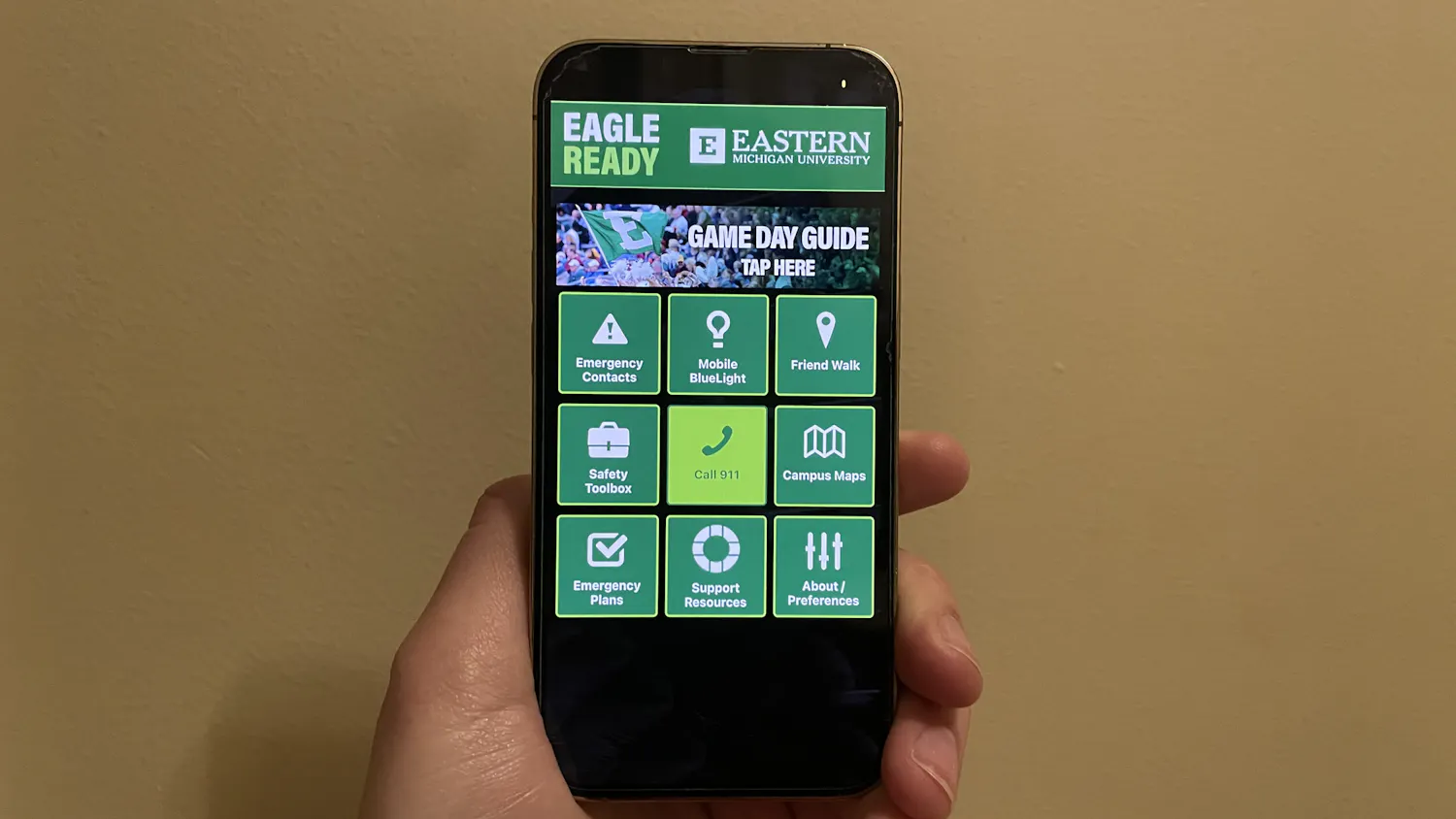 A hand holding a phone that is displaying a black and green app. The home page has buttons for various emergency services and campus resources.