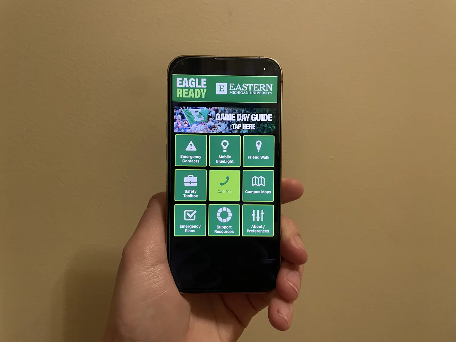 A hand holding a phone that is displaying a black and green app. The home page has buttons for various emergency services and campus resources.