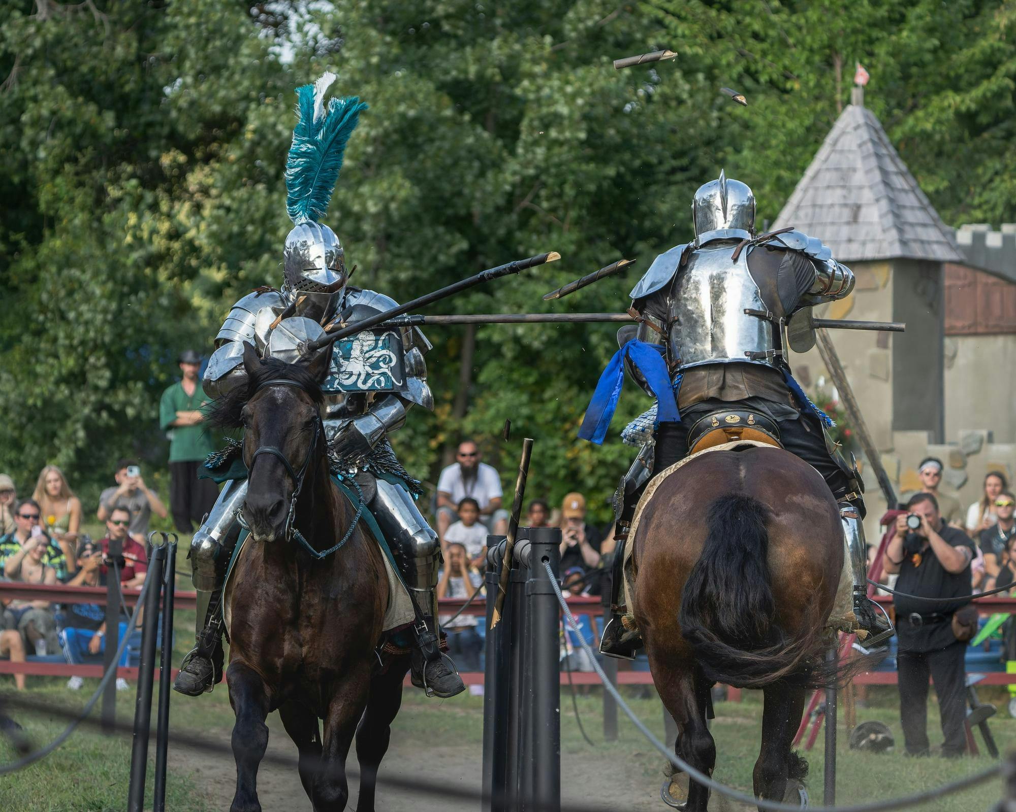 Two actors dressed as armored knights ride brown horses past each other while jousting with long sticks.