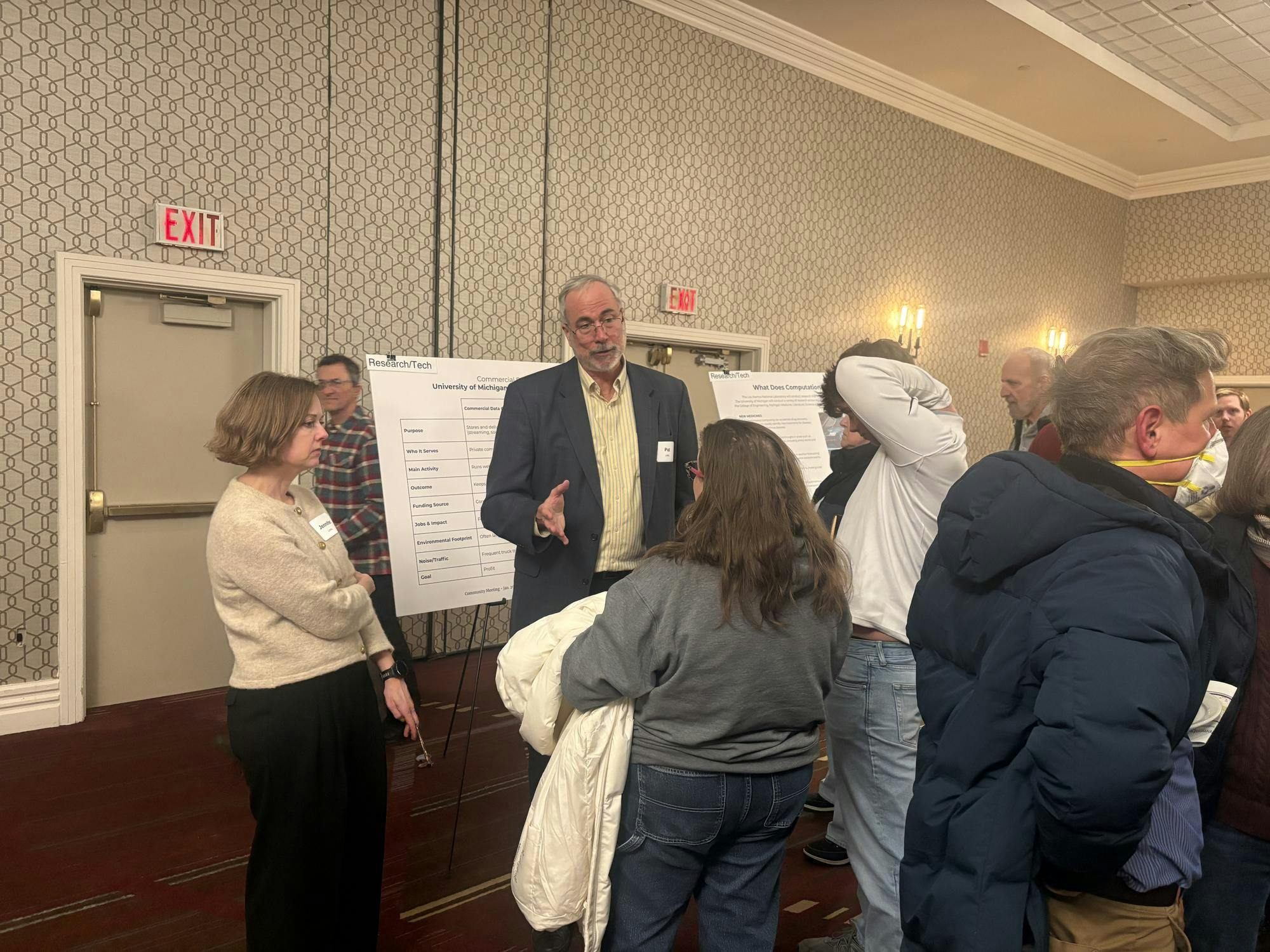 Image of Patrick Fitch addressing a group of people in a hotel conference room. In the background, there are poster boards presenting information about the project and the walls are beige with a hexagon pattern design. 