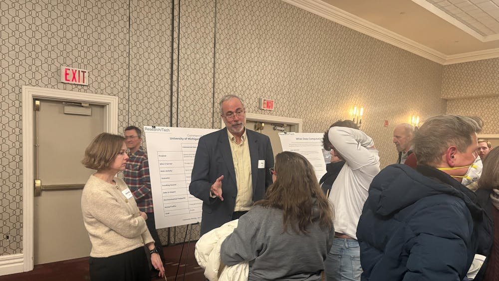 Image of Patrick Fitch addressing a group of people in a hotel conference room. In the background, there are poster boards presenting information about the project and the walls are beige with a hexagon pattern design.