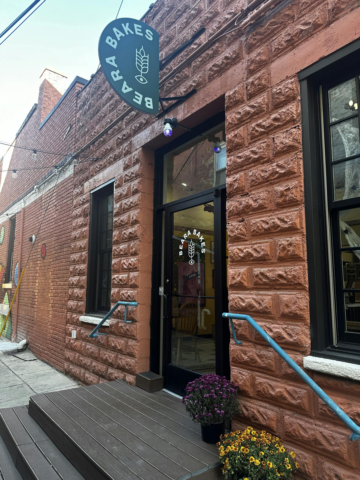 A brick building has a glass door with the words Beara Bakes in white and a black sign above it with the words Beara Bakes in white. 