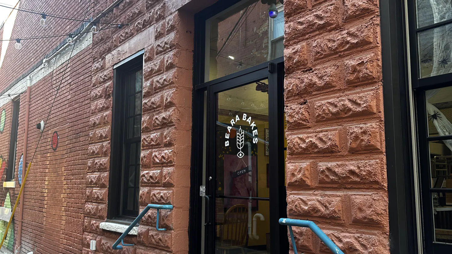 A brick building has a glass door with the words Beara Bakes in white and a black sign above it with the words Beara Bakes in white.